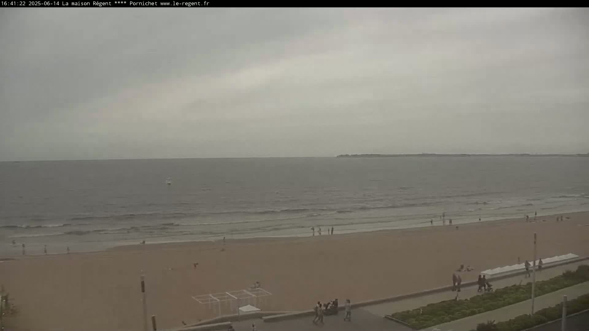 A wide shot shows a sandy beach with a few people scattered across it, gentle waves lapping the shore under an overcast sky.