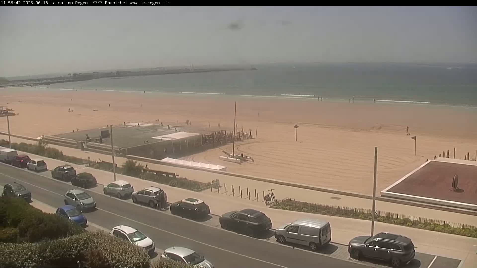 A sandy beach with a few people on it and cars parked along a road next to the beach on a hazy sunny day.