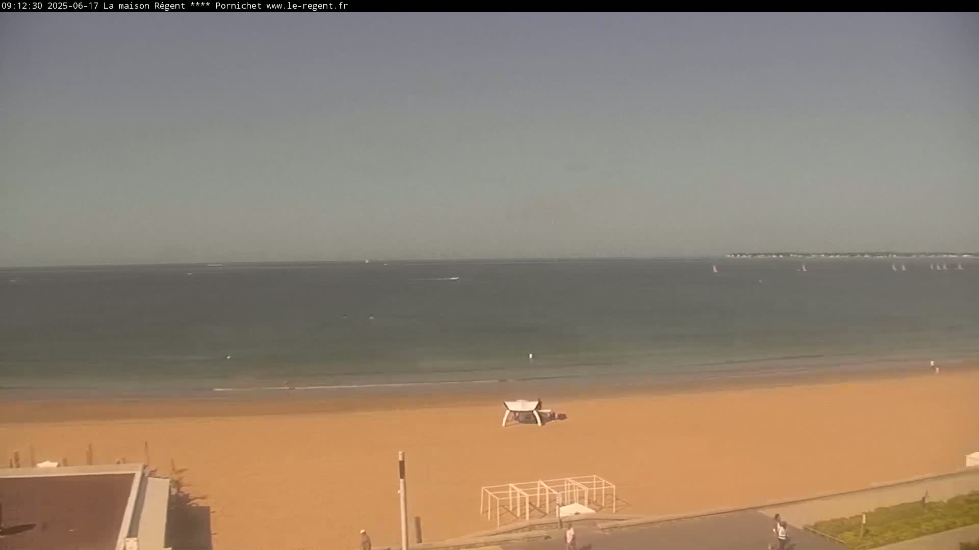 A mostly sunny day at a beach with calm water, a few sailboats in the distance, and a sandy shore with some beach structures.