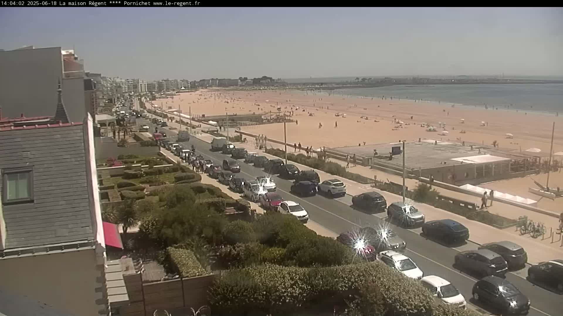 A sunny day at a beach shows many people sunbathing and swimming, with a road lined with parked cars alongside a building and lush greenery.