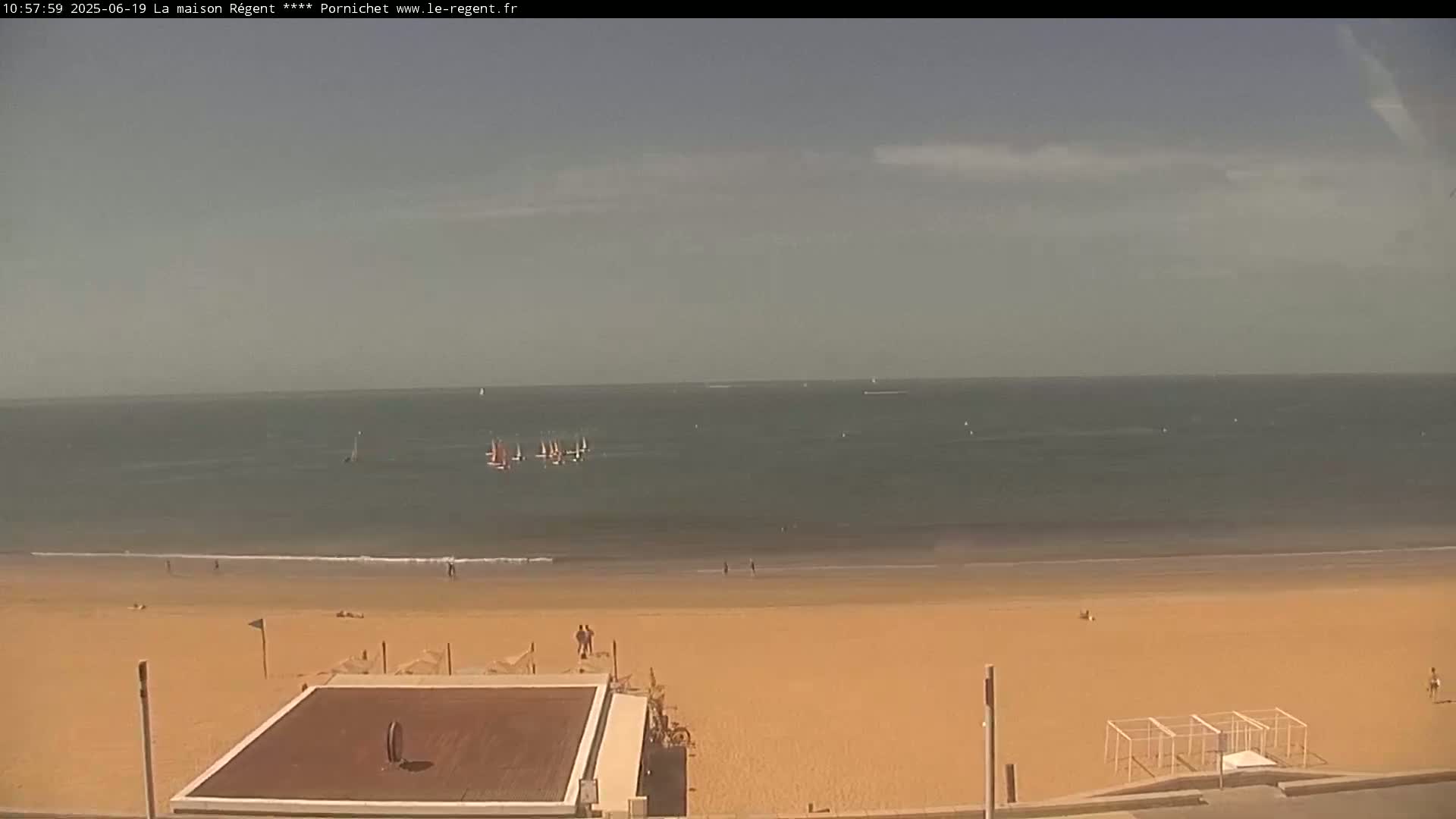 A mostly sunny day shows a sandy beach with several people, a dark-colored square structure, and sailboats visible in the ocean.