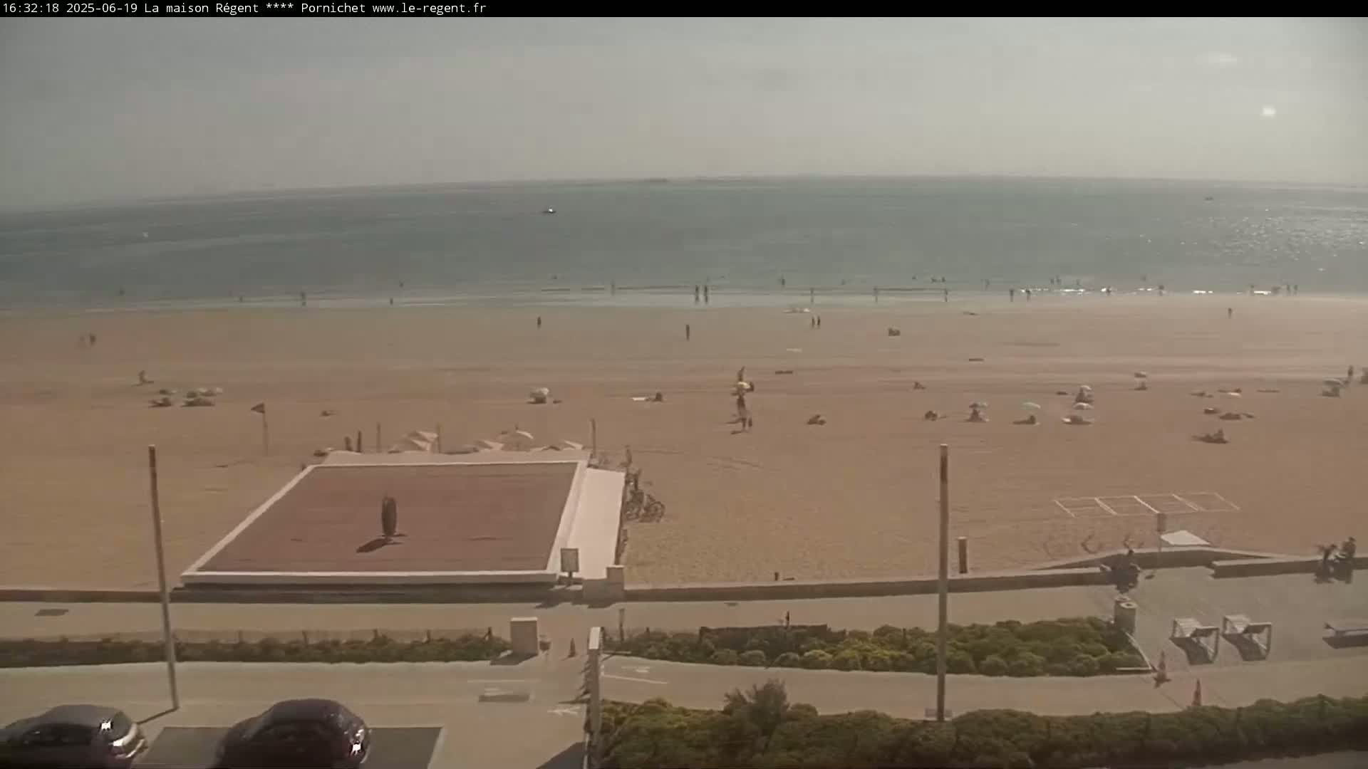 A sandy beach with scattered sunbathers and a volleyball court is seen from an elevated viewpoint on a hazy, sunny day, with a calm ocean in the background.