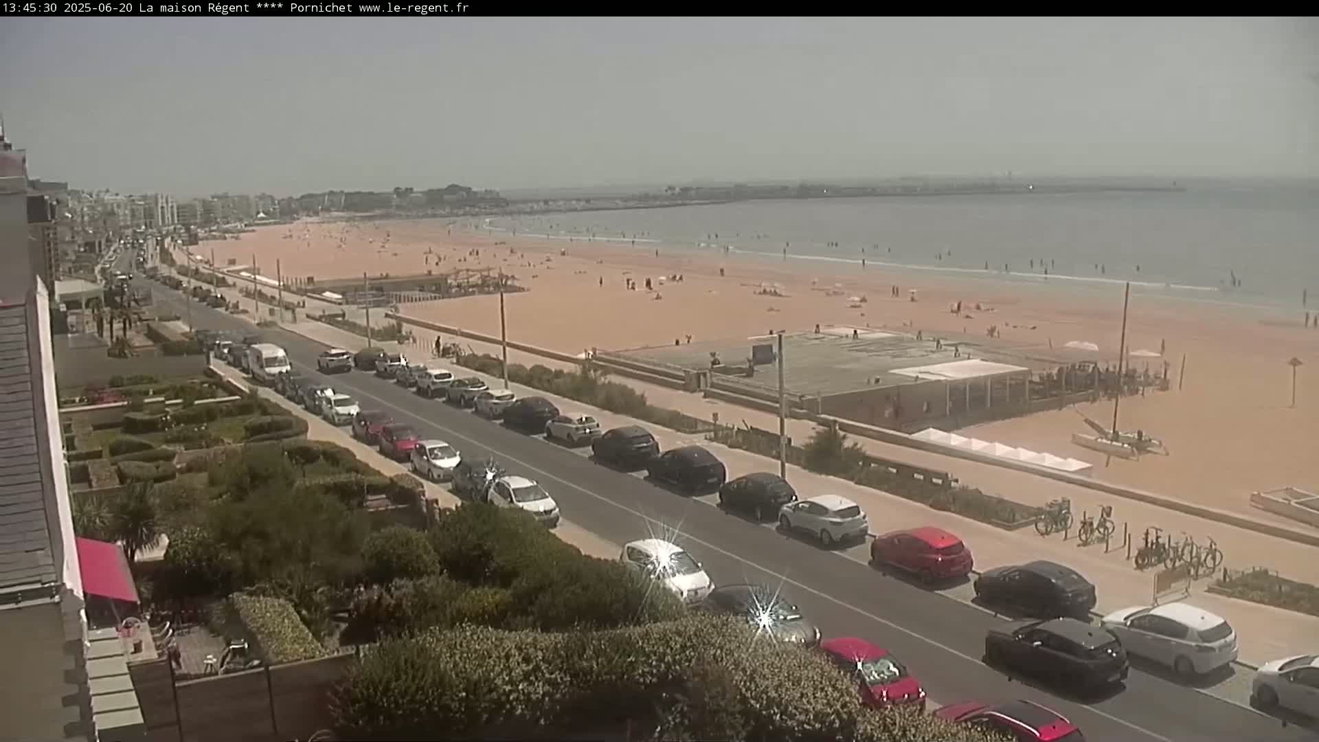A sandy beach with people sunbathing and swimming is visible from a high vantage point, showing a street lined with parked cars and buildings beyond, under a hazy sky.