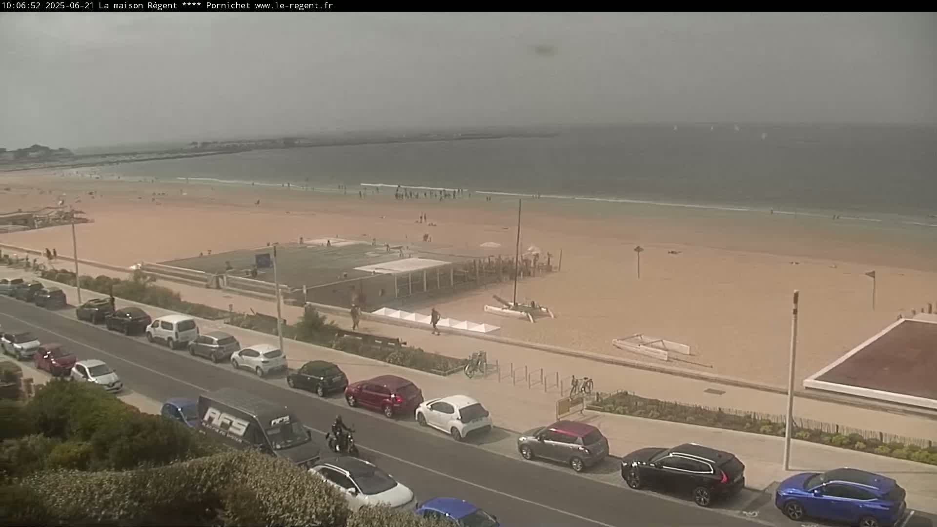 A hazy beach scene shows a sandy shore with scattered people, a few parked sailboats, and a roadway lined with parked cars.