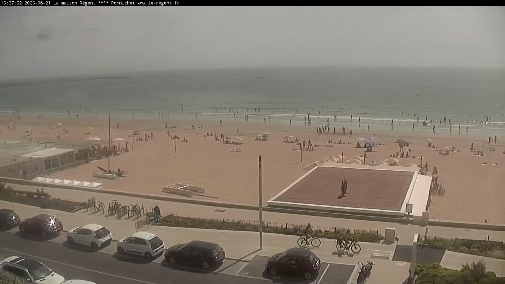 A hazy, sunny day at a sandy beach with many people swimming, sunbathing, and walking, with cars parked along a nearby road and bicycles leaning against a fence.