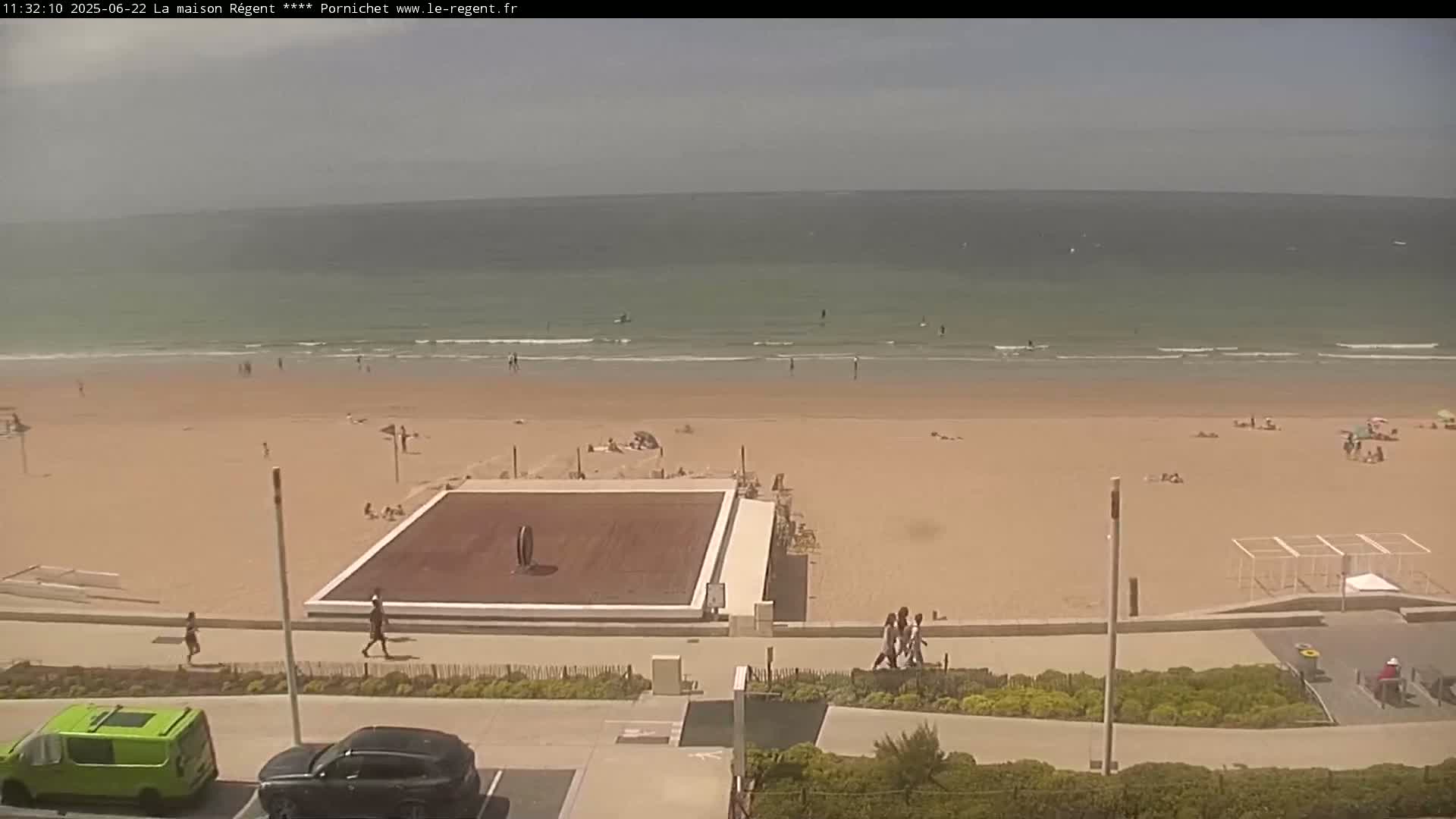 An overhead view shows a sandy beach with people sunbathing and swimming in a calm ocean under a partly cloudy sky.
