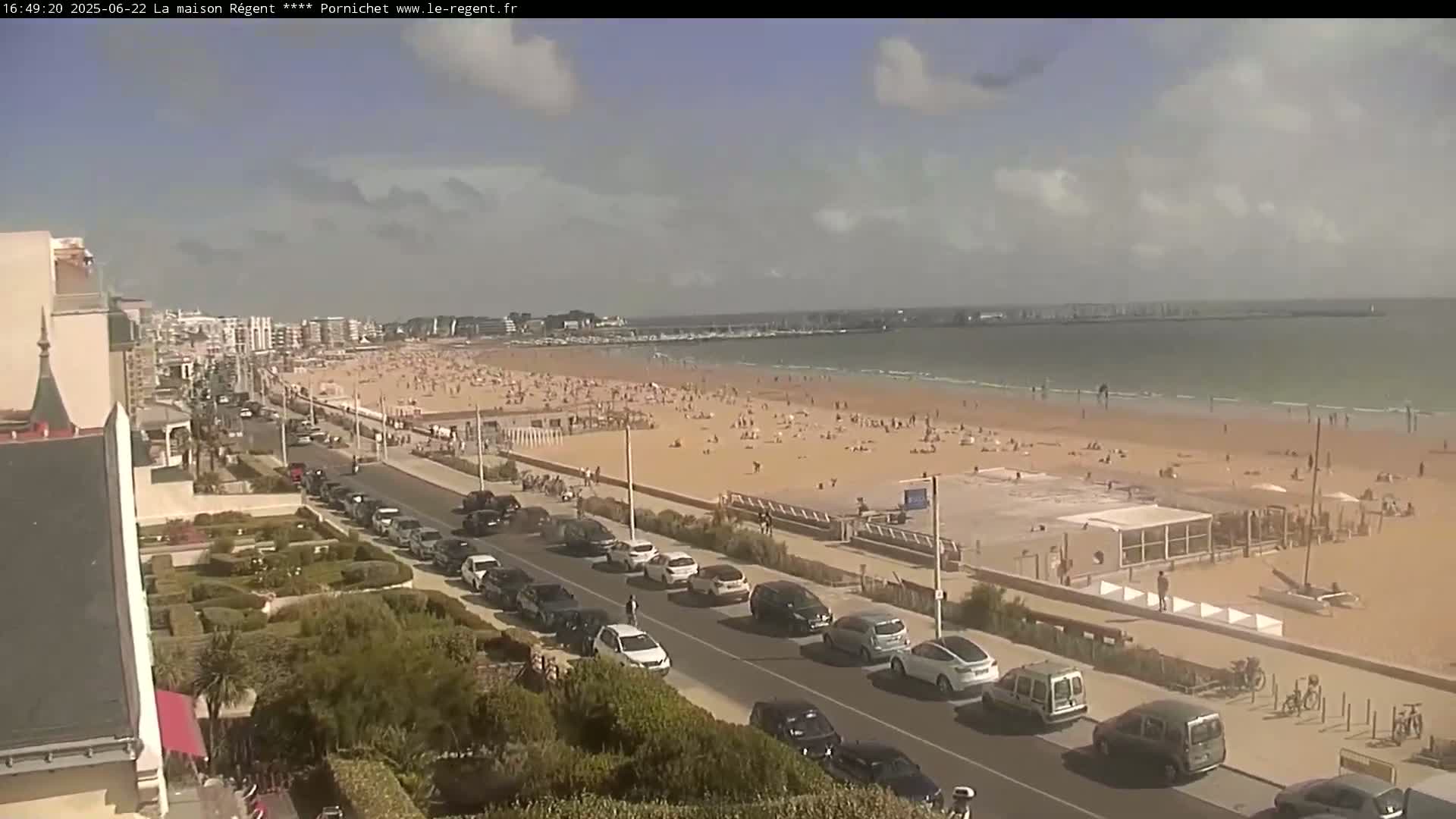 A sunny day at a crowded beach with a roadway and parked cars lining the shore, viewed from an elevated position.