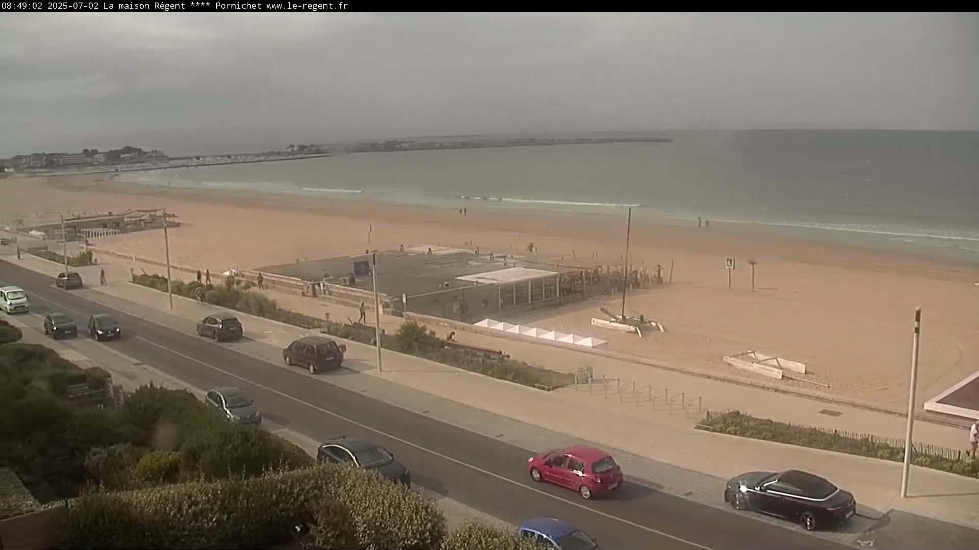 A sandy beach with calm ocean waves and several cars parked along a roadside next to a beachfront promenade on an overcast day.