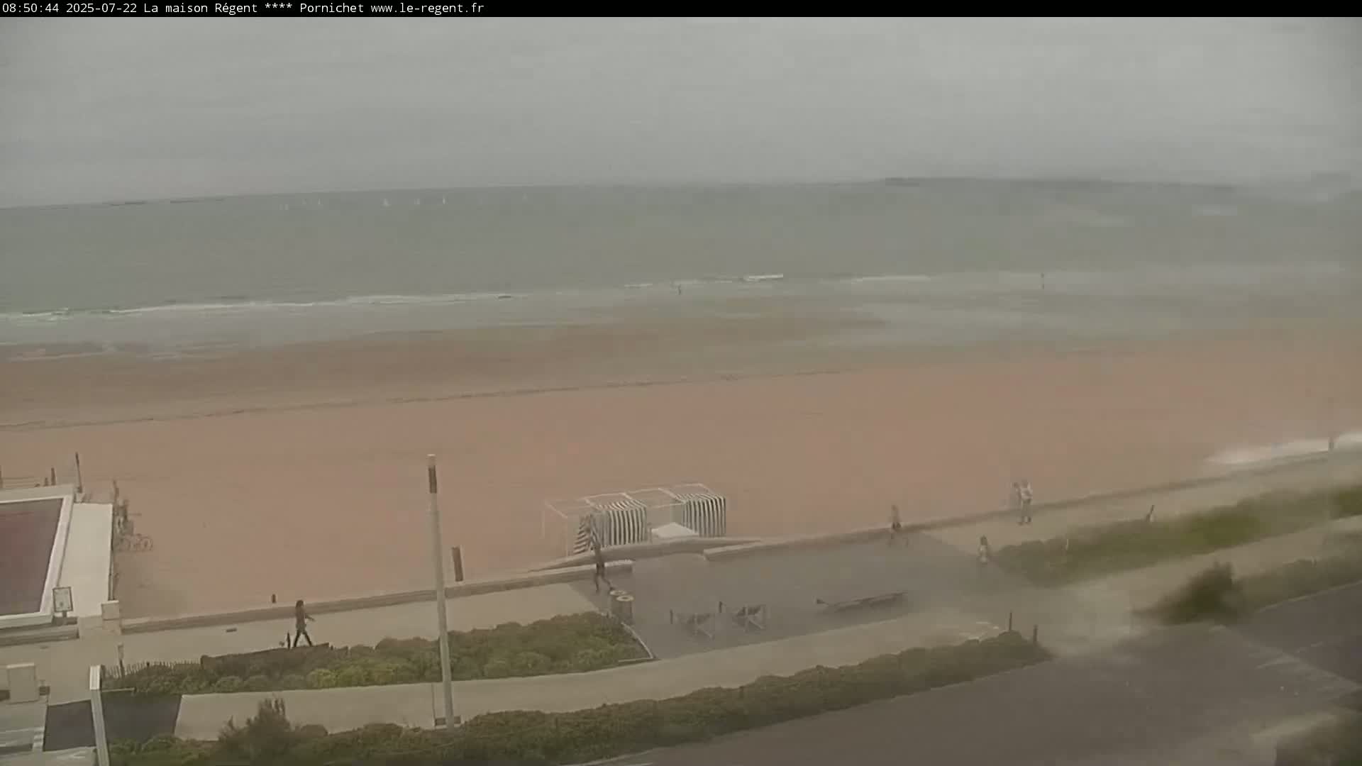 A wide shot shows a beach with several people walking along the boardwalk on an overcast day with choppy waves.