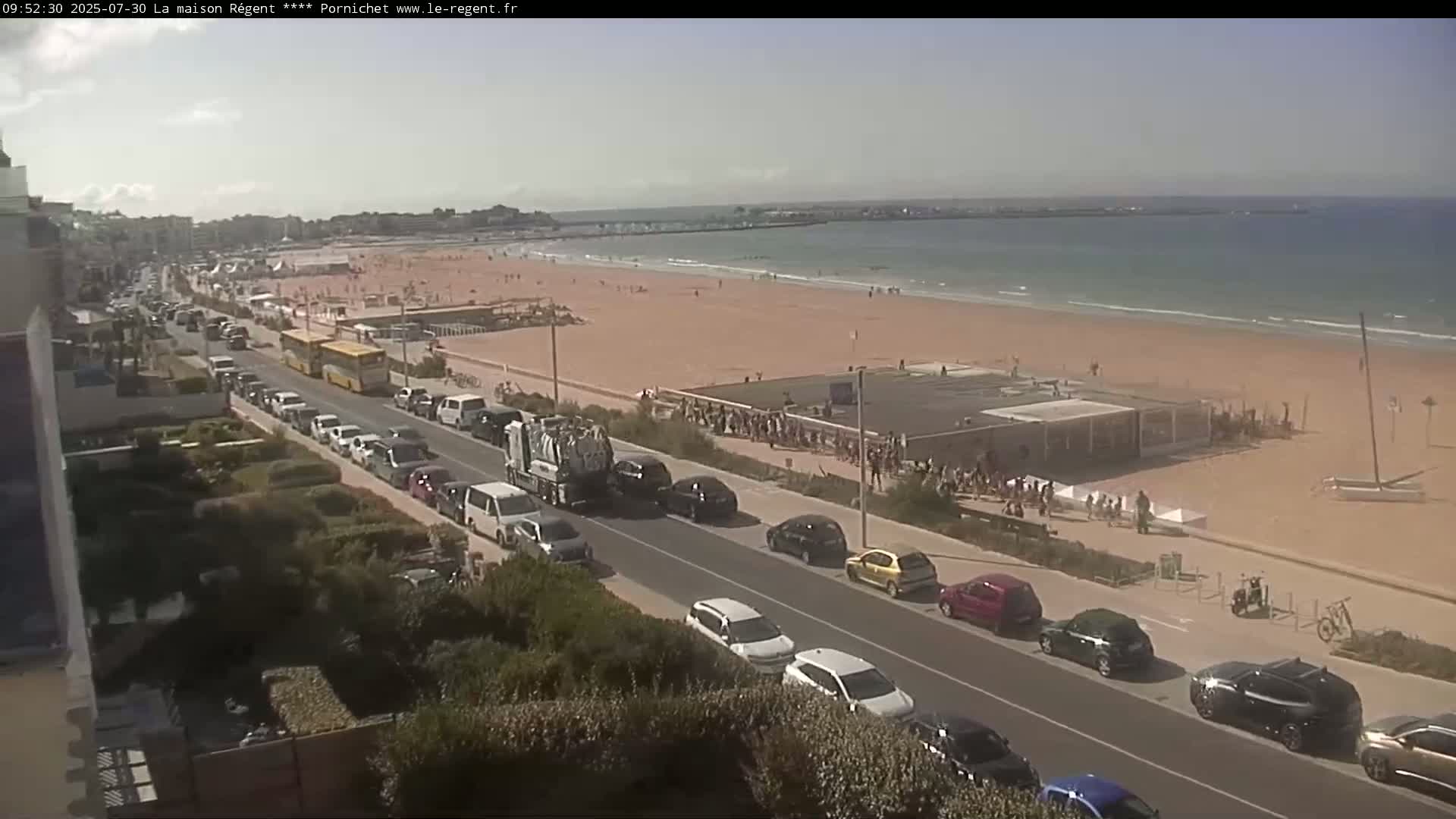 A sunny day at a beach with many people on the sand and a road lined with cars leading away from the shore.