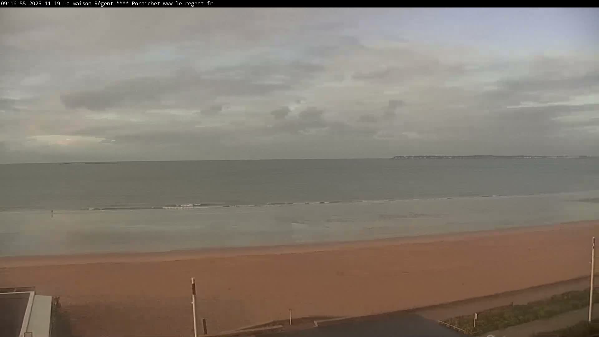 A wide view captures a sandy beach, a grey ocean with small waves, and a distant coastline under a heavily overcast sky.