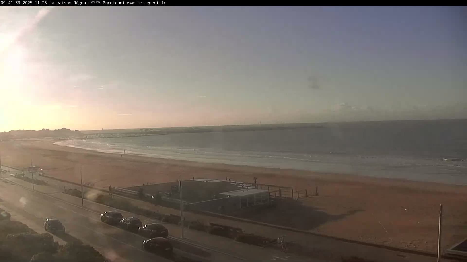 A wide sandy beach stretches along a calm sea under a bright, clear sky with low morning sun, featuring a parallel road with cars, beachside structures, and a few people walking.