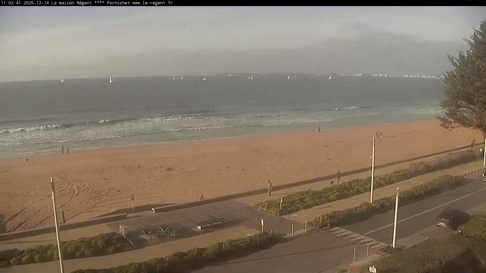 A wide sandy beach stretches along a calm sea under a bright, clear sky with low morning sun, featuring a parallel road with cars, beachside structures, and a few people walking.
