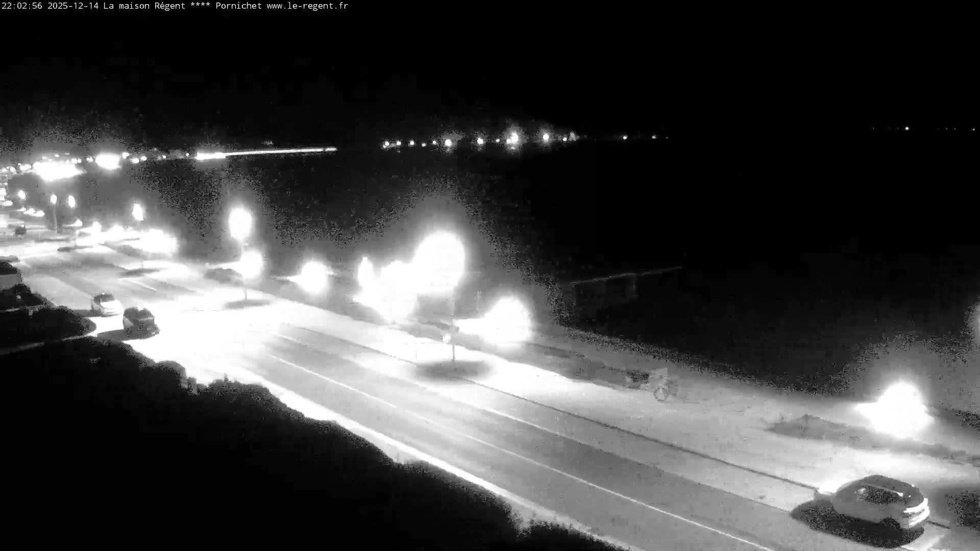 A wide sandy beach stretches along a calm sea under a bright, clear sky with low morning sun, featuring a parallel road with cars, beachside structures, and a few people walking.