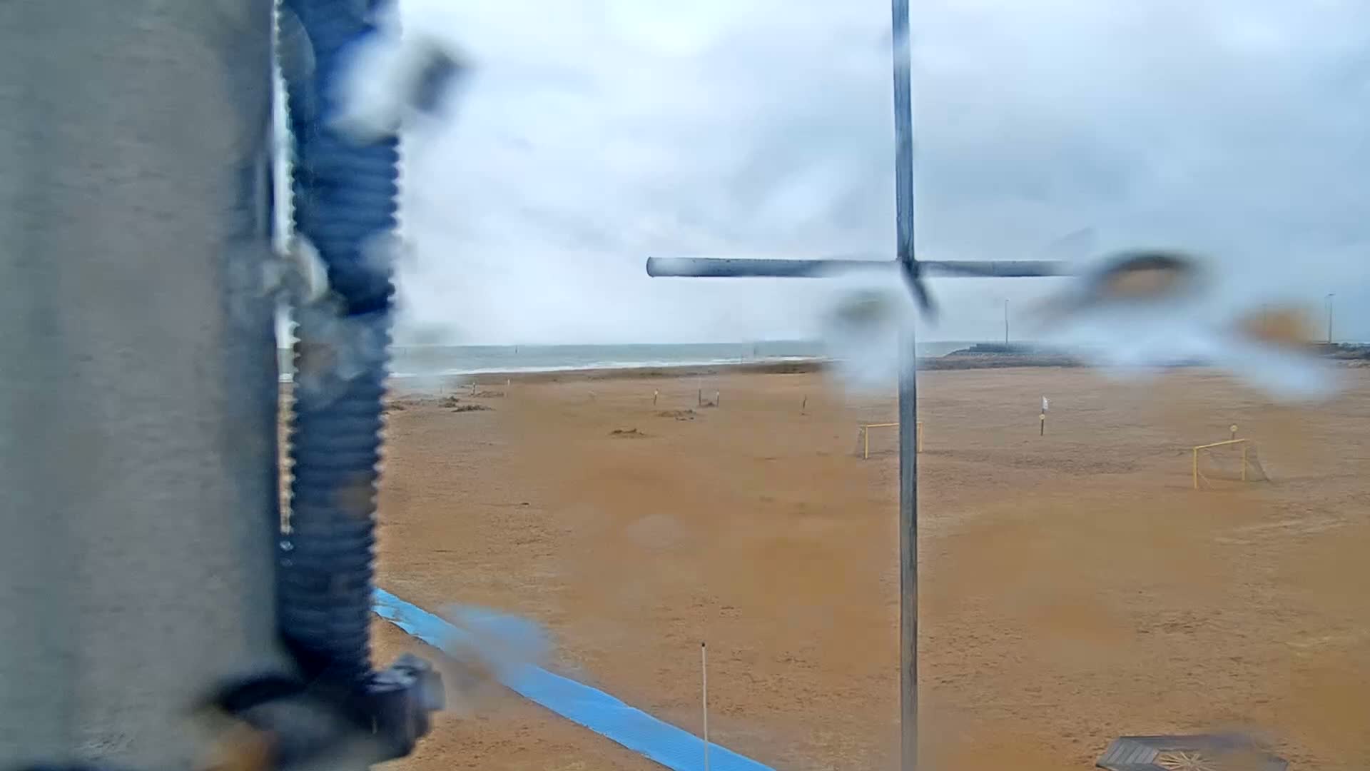 A rain-covered view shows a sandy beach with soccer goals, a wavy ocean in the distance, and a metal pole in the foreground.