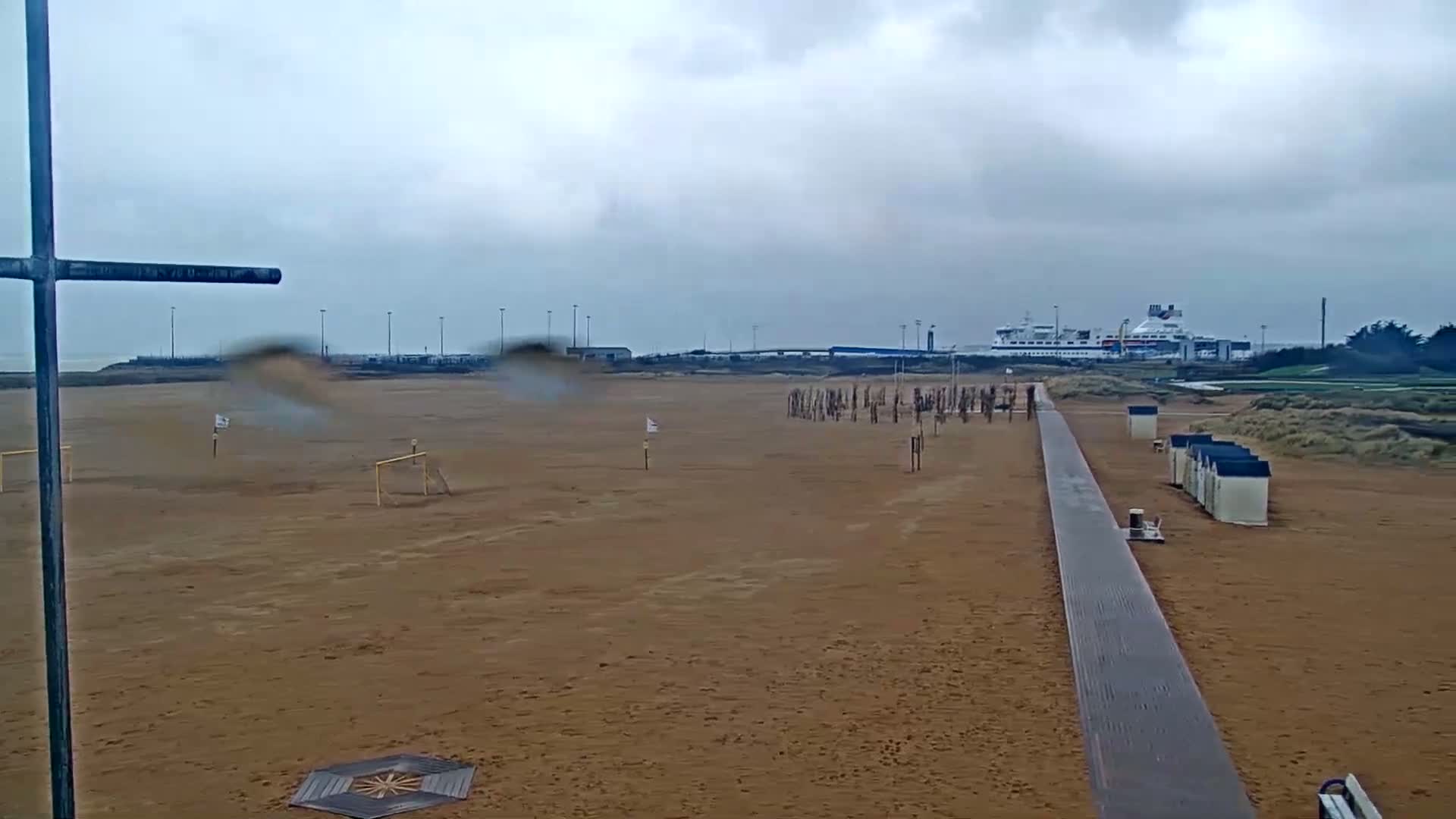 A sandy beach with a boardwalk, soccer goals, beach huts, and a large ferry visible in the distance under an overcast sky with light rain.