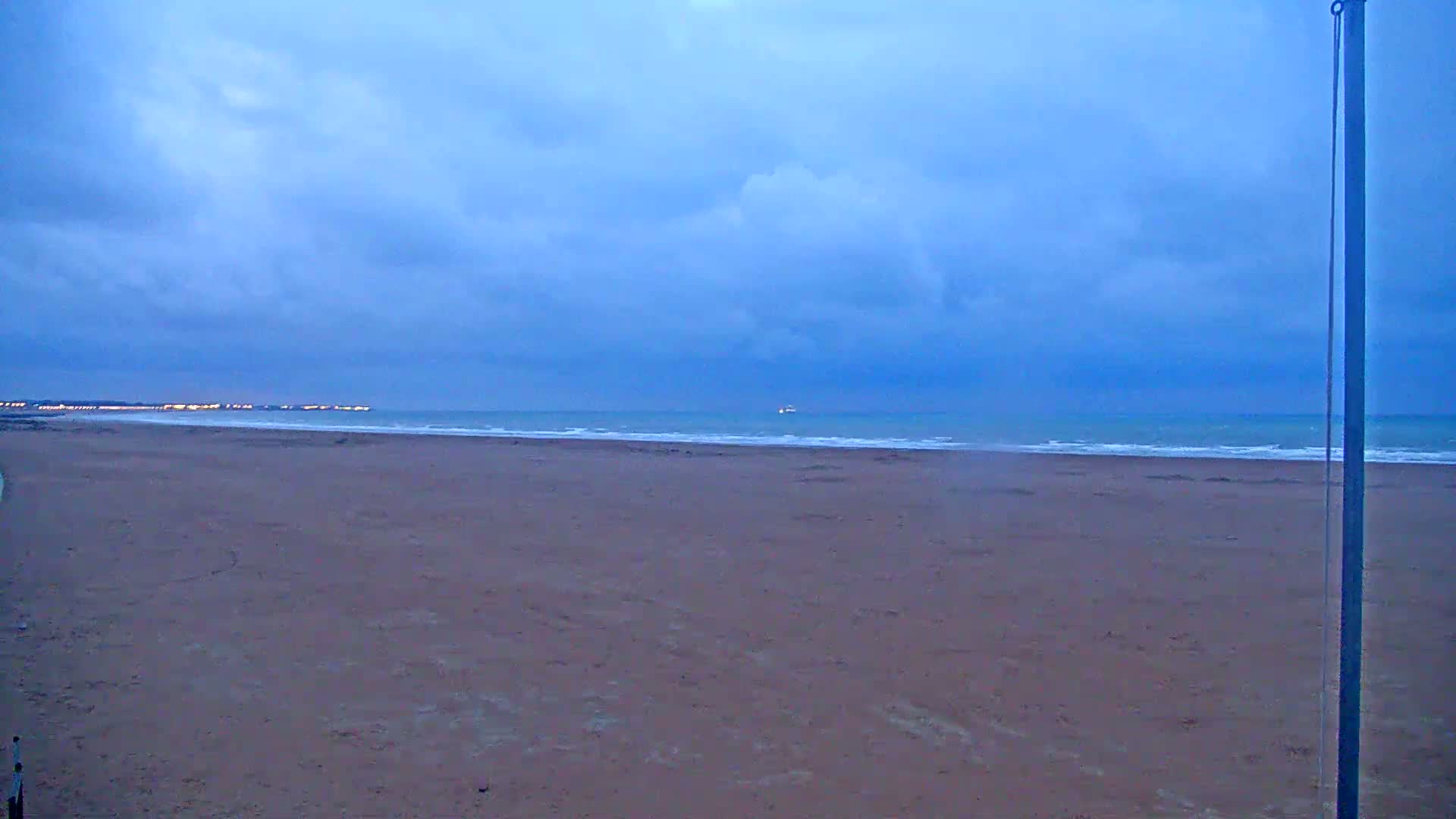 A wide, flat sandy beach stretches to a calm ocean under a mostly cloudy, dusky blue sky.