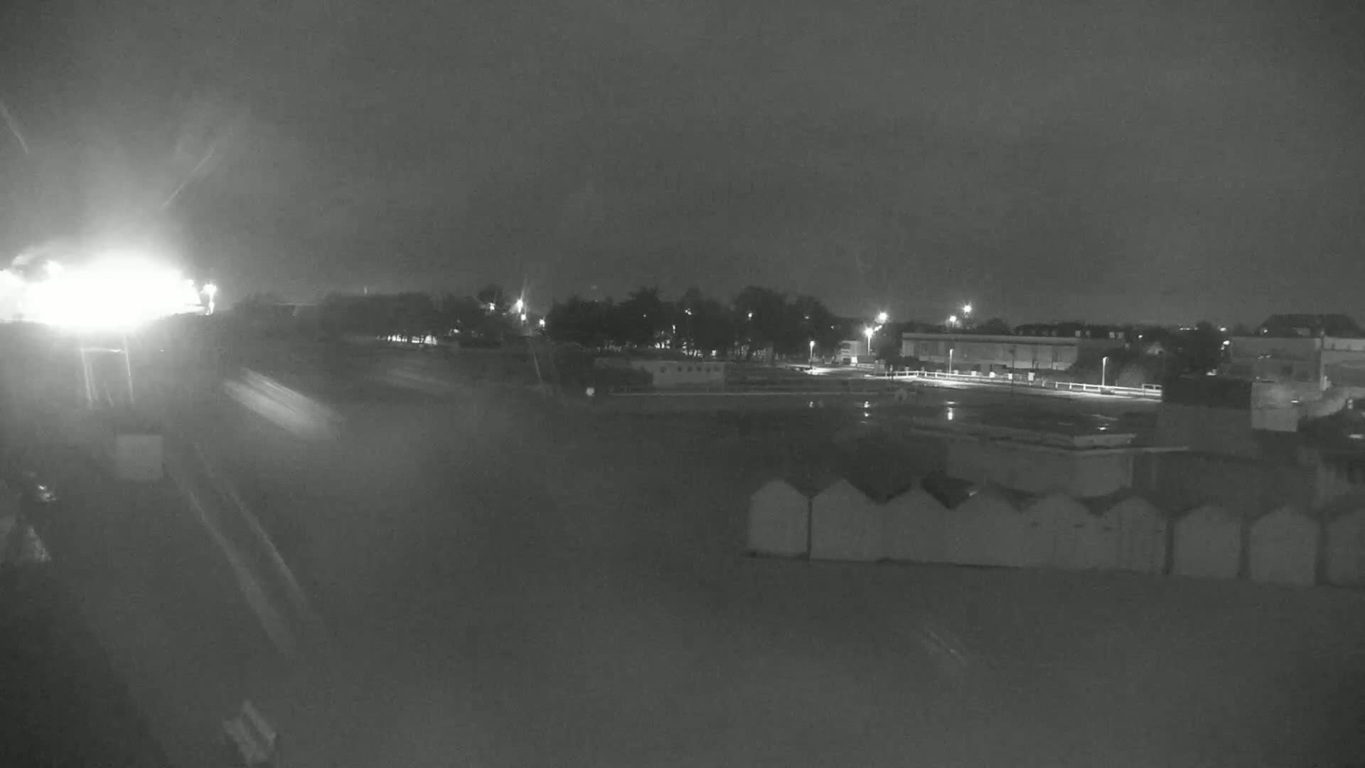 A nighttime, possibly rainy, view of a beach area with rows of beach huts and buildings in the distance, illuminated by streetlights.