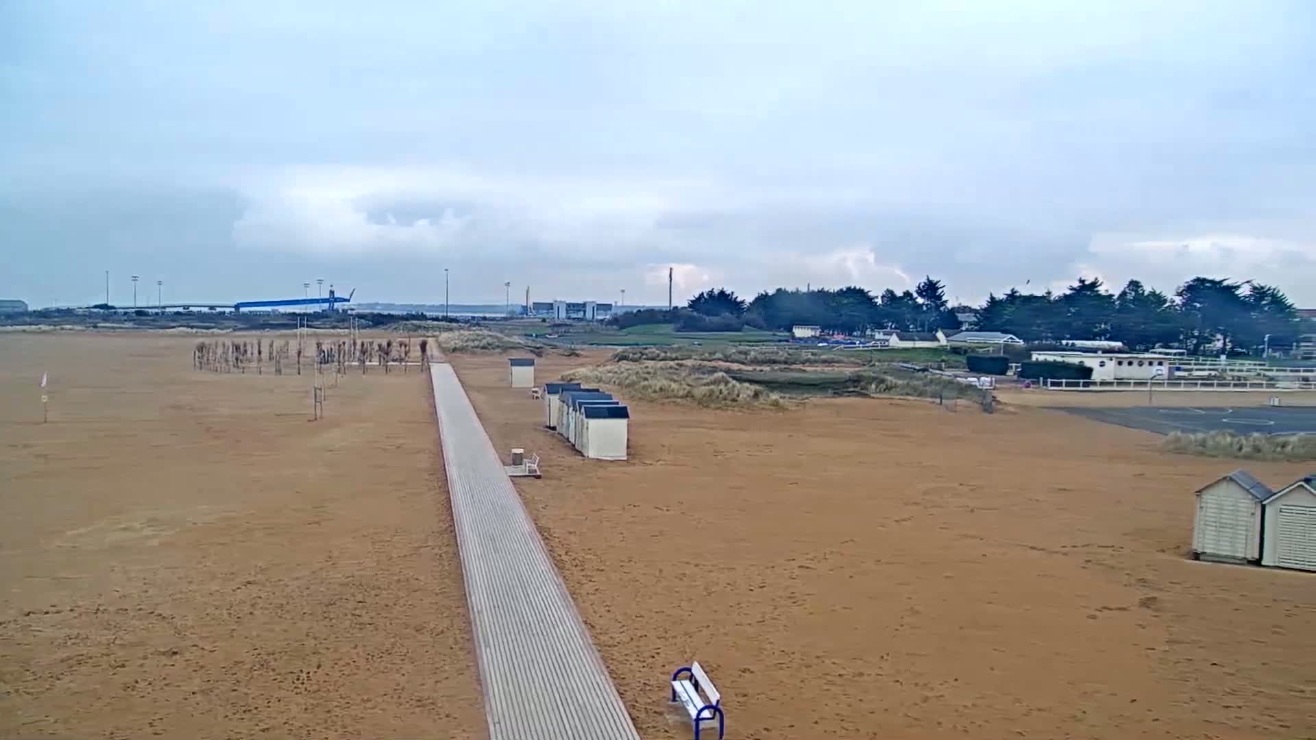 A sandy beach with a boardwalk, beach huts, and a distant line of trees under a cloudy sky.