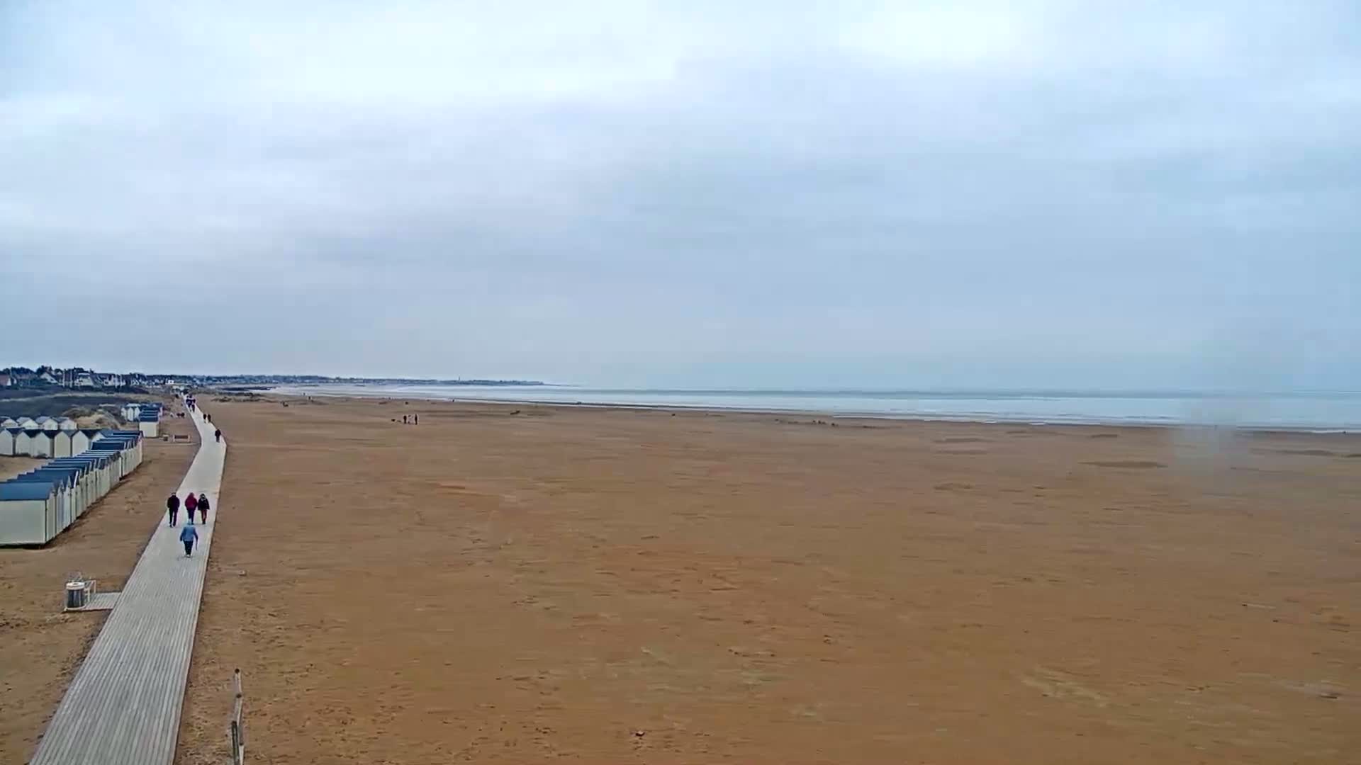 A long, wide sandy beach stretches to a calm, gray ocean under an overcast sky, with a boardwalk and several people walking along it.