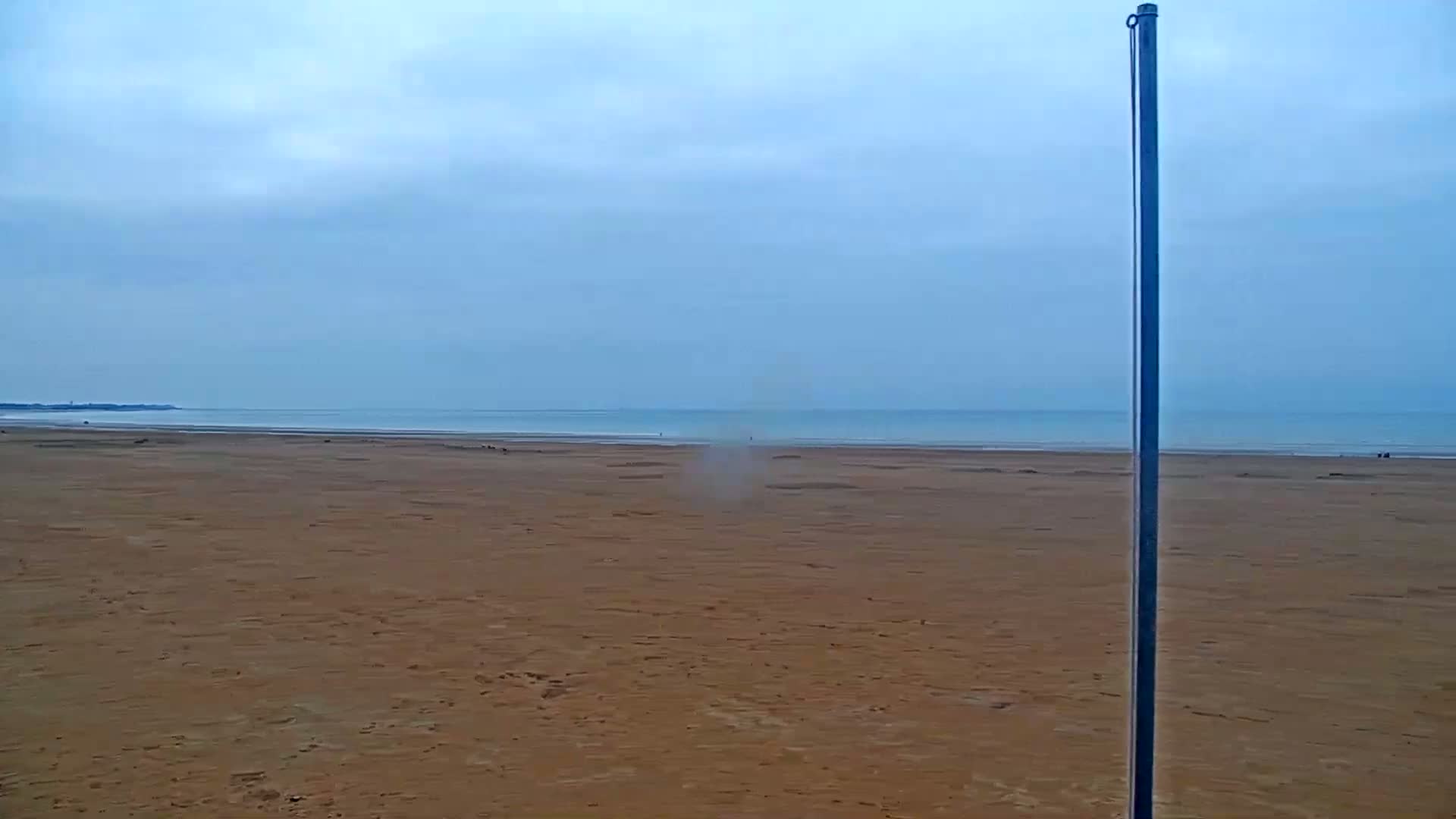 A tall pole stands on a wide, flat, sandy beach under a cloudy sky, with a calm sea in the distance.