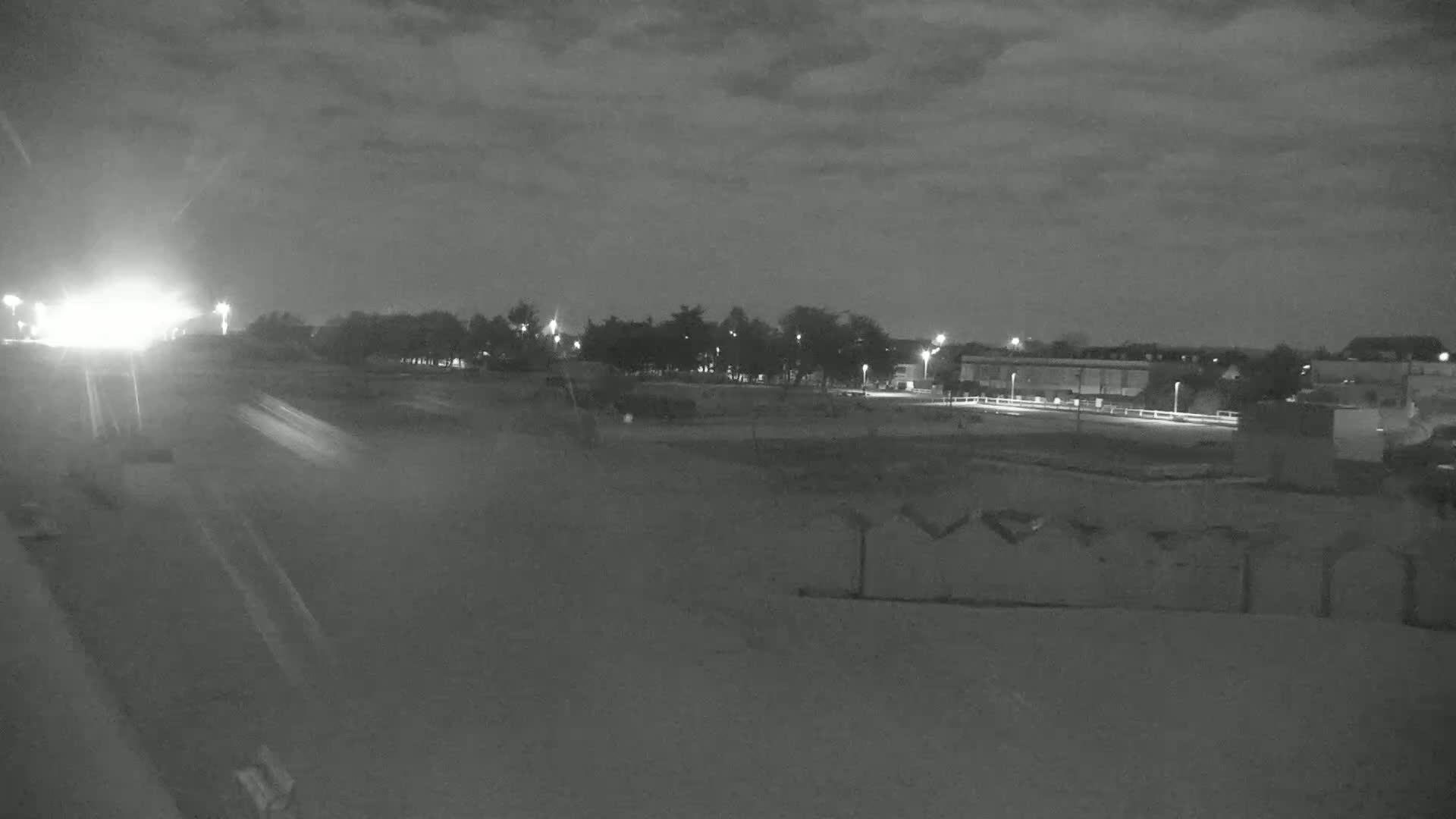 A nighttime, overcast scene shows a road with headlights in the distance, leading to a line of trees and buildings under dimly lit streetlights.