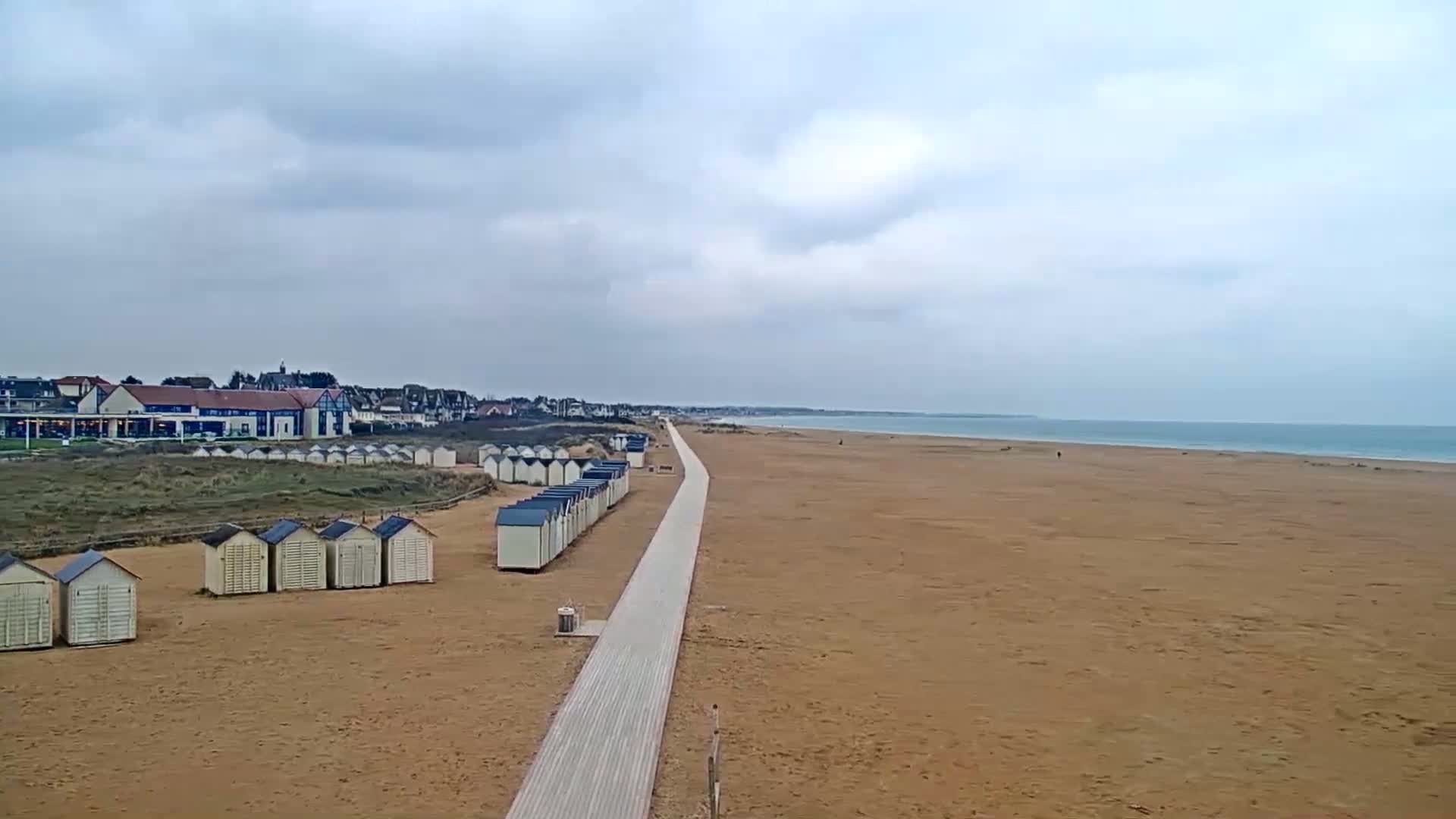 A long, sandy beach stretches to a calm ocean under an overcast sky, with a boardwalk leading from a row of beach huts towards the water.