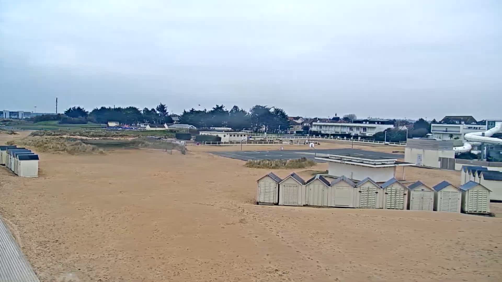 A sandy beach with rows of beach huts, an outdoor sports court, and buildings in the background under an overcast sky.