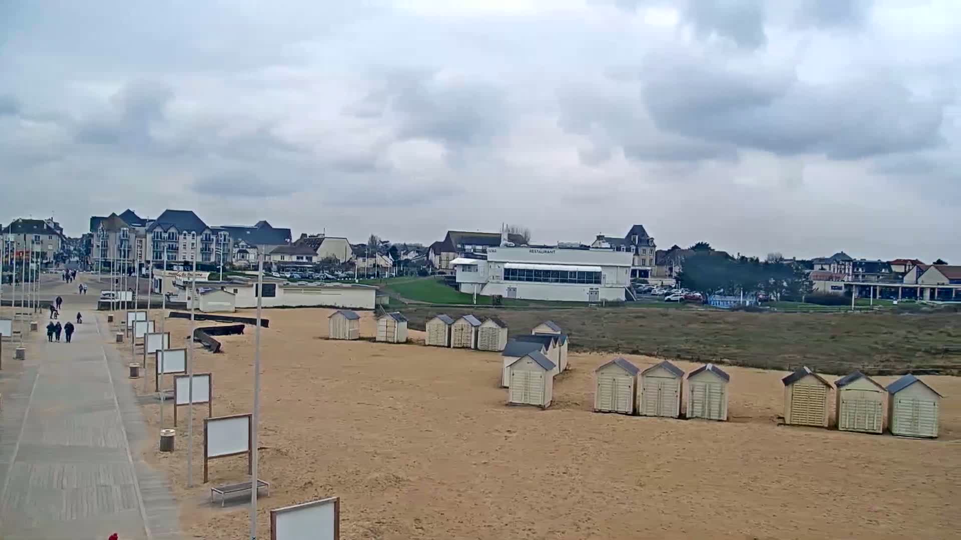 A sandy beach with rows of small beach huts, a boardwalk with people walking, and buildings in the background under a cloudy sky.