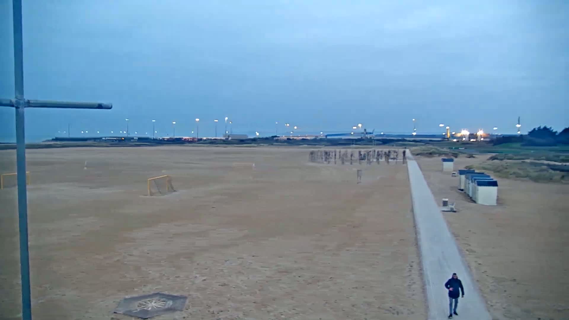 A person walks down a paved path on a sandy beach at dusk, near some beach huts and soccer goals, with a distant industrial area faintly visible under a clear, twilight sky.