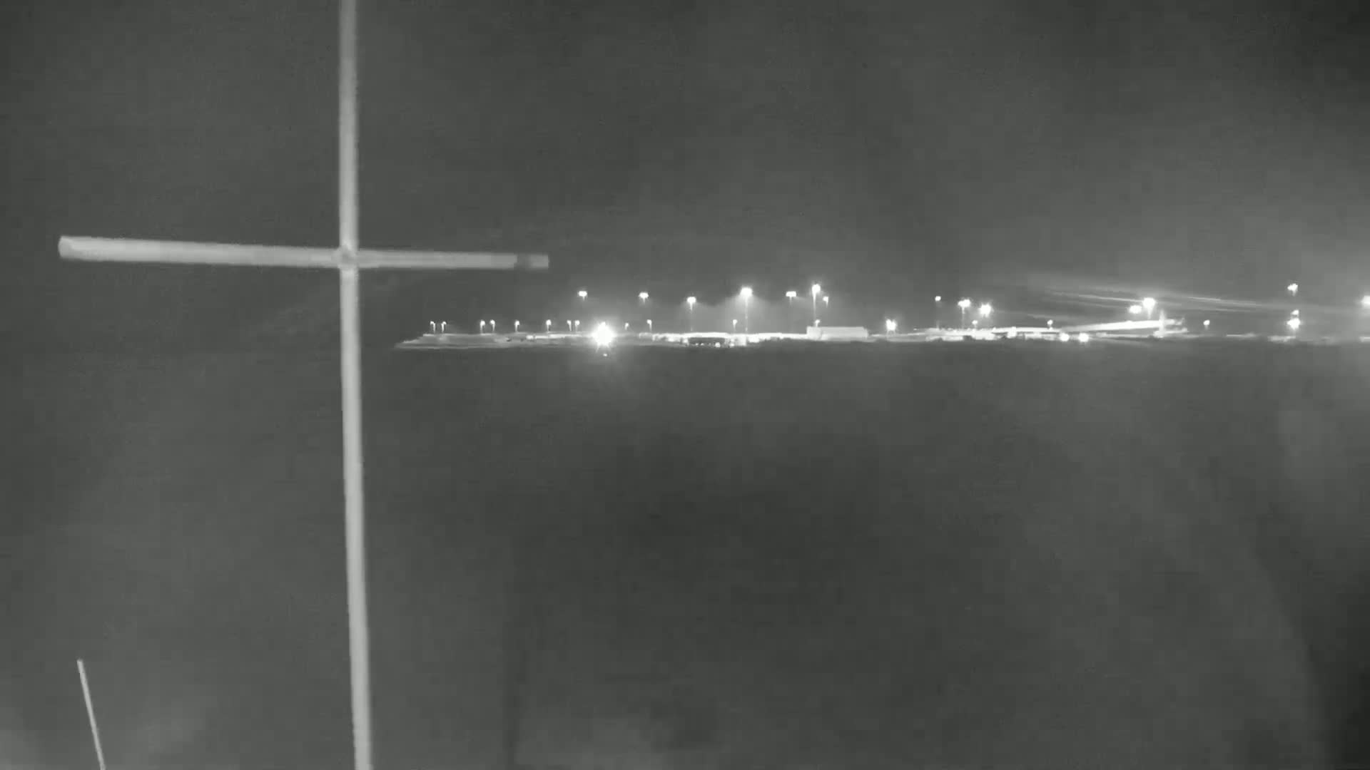 A large cross stands in the foreground against a nighttime background of dimly lit buildings and land.