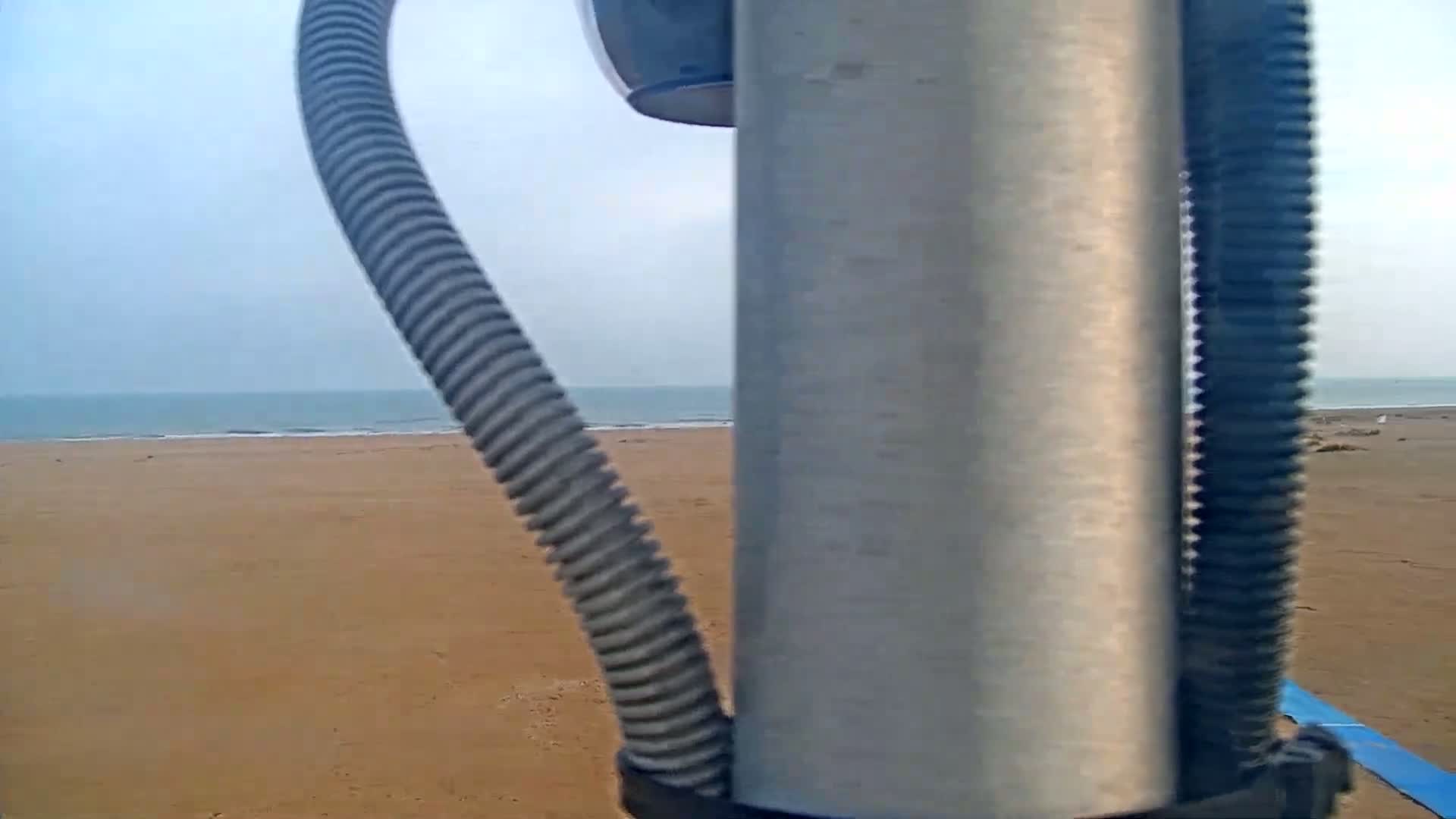 A view from behind a corrugated tube and metal pole shows a calm ocean and a sandy beach under a mostly clear sky.
