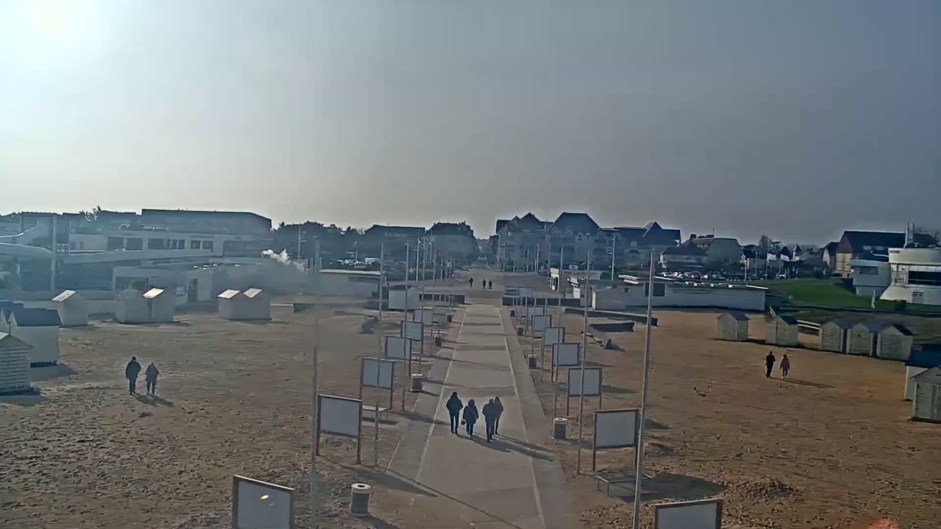A paved walkway leads from a sandy beach toward a row of buildings under a hazy, sunny sky.