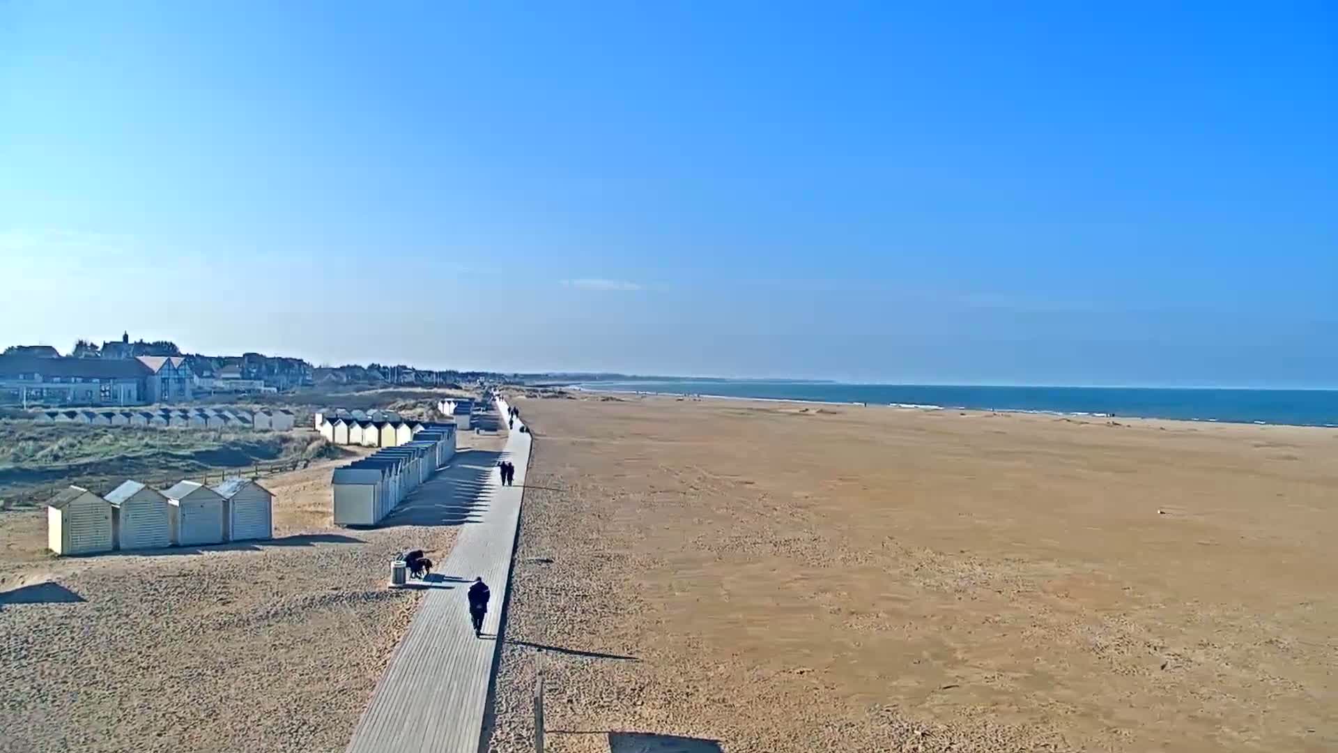 A boardwalk runs along a wide, sandy beach toward a calm ocean under a clear blue sky.