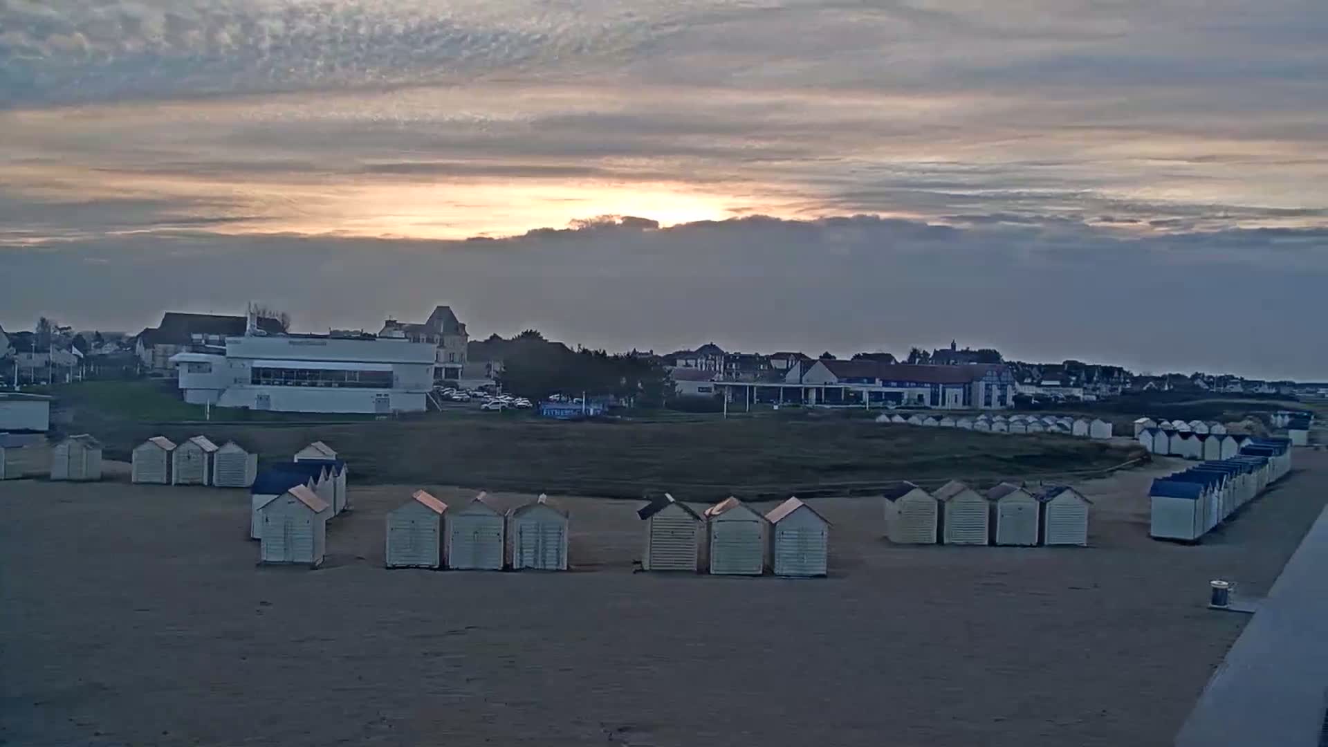 Rows of beach huts line a sandy beach at sunset under a mostly cloudy sky.