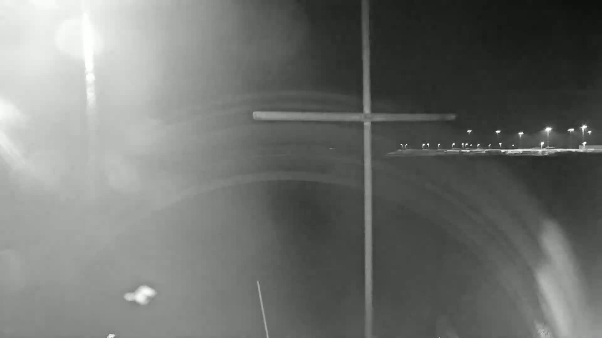 A large cross stands against a night sky with bright lights in the distance and hazy, blurry light sources in the foreground.