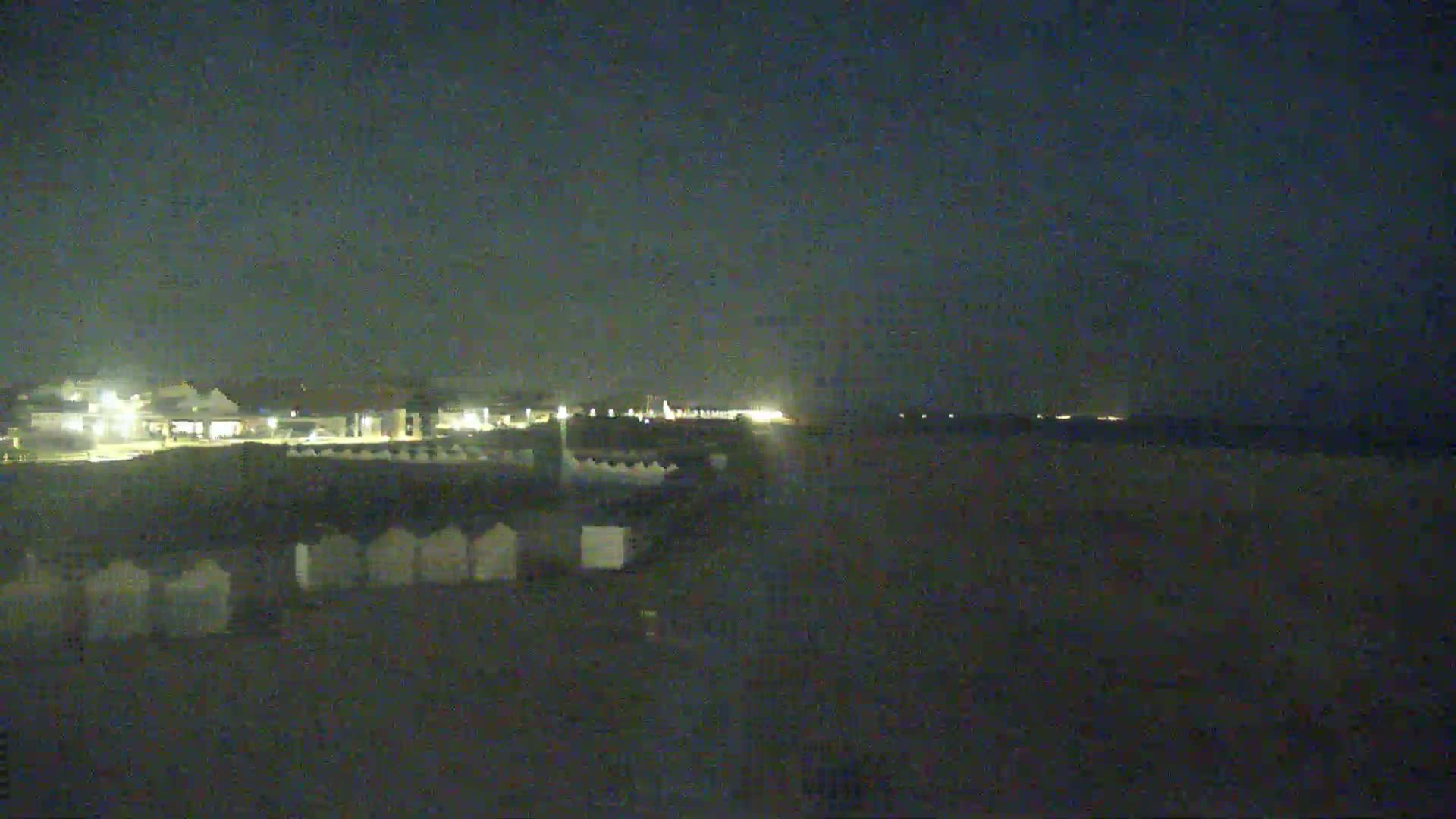 A dark, nighttime beach scene shows a row of beach huts and a dimly lit shoreline with a body of water beyond.