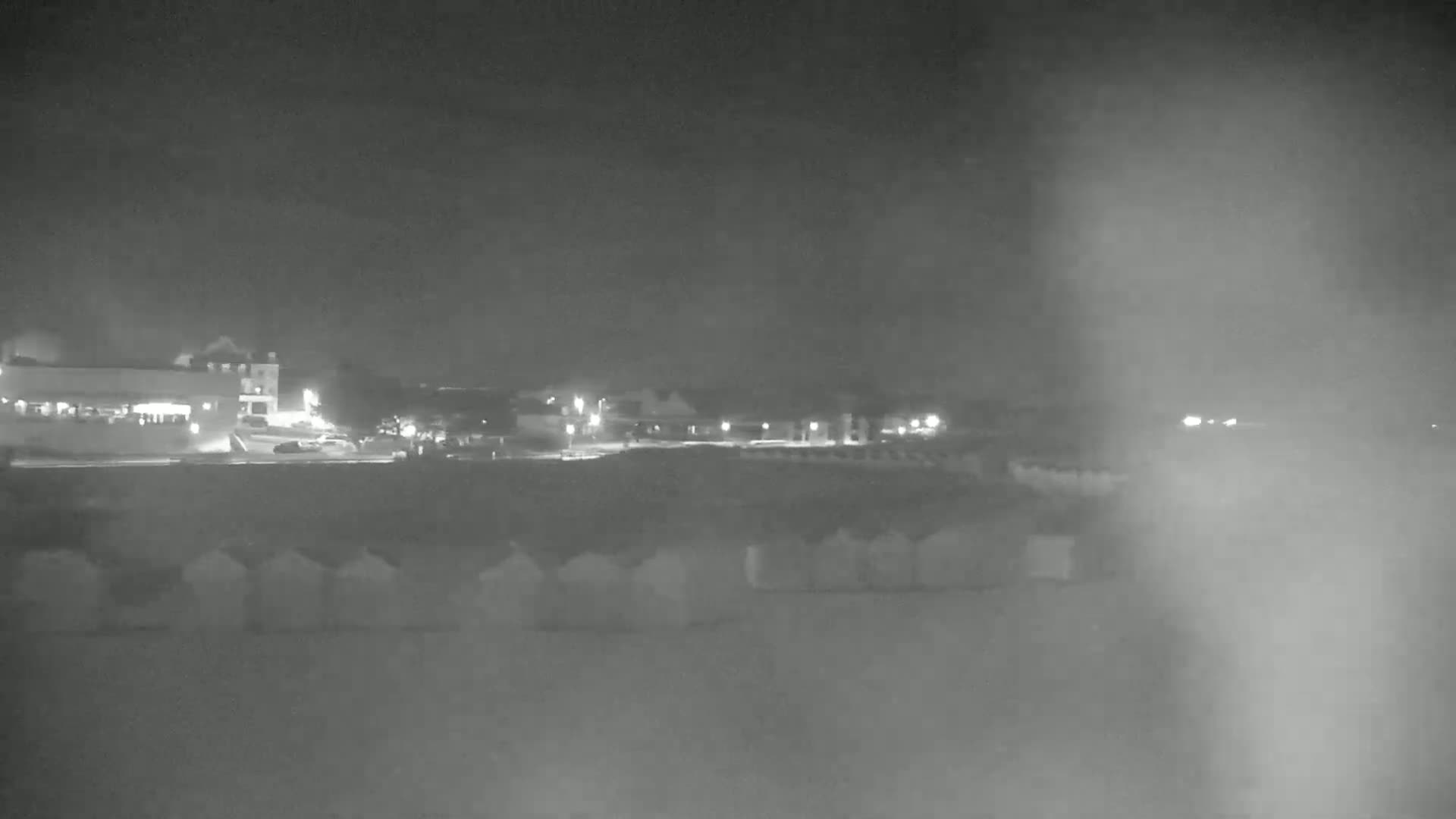 A nighttime, hazy view shows a row of beach huts in front of a dimly lit shoreline with buildings and some parked cars visible in the distance.