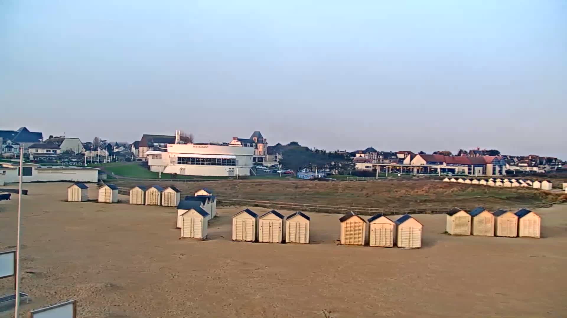 A sandy beach with rows of small beach huts, backed by buildings and a grassy area, under a clear, light sky.