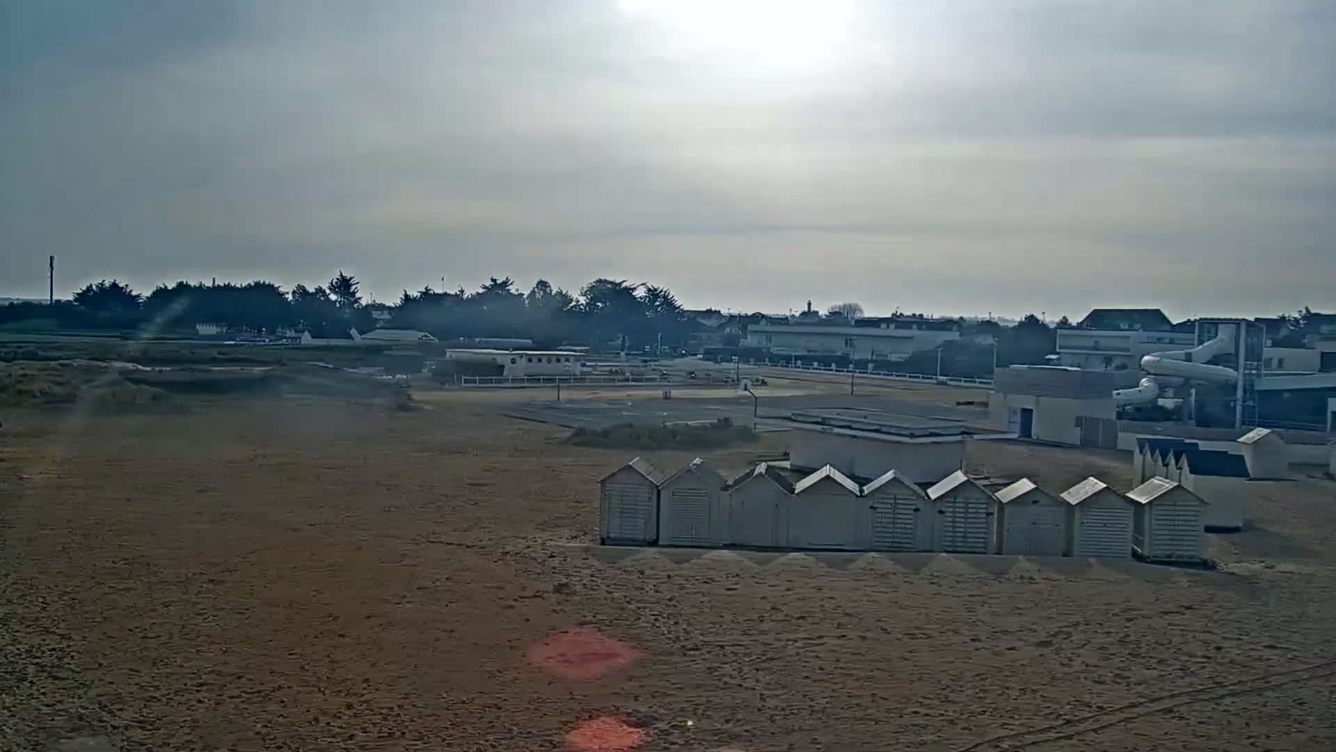 A sandy beach with beach huts, a basketball court, and buildings in the background under a bright, overcast sky.