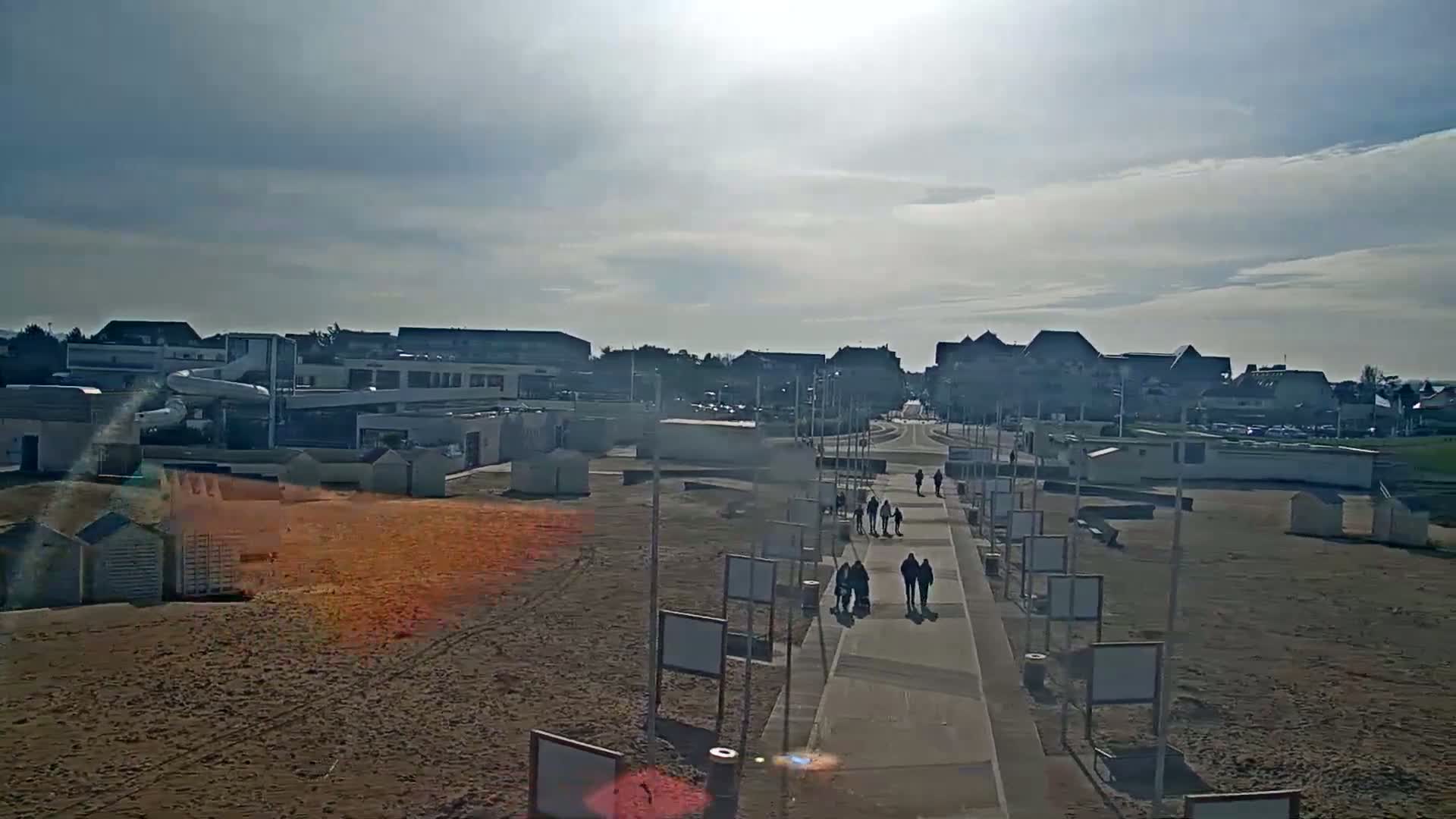 A paved walkway leads to a sandy beach with beach huts and buildings in the background under a partly sunny sky.
