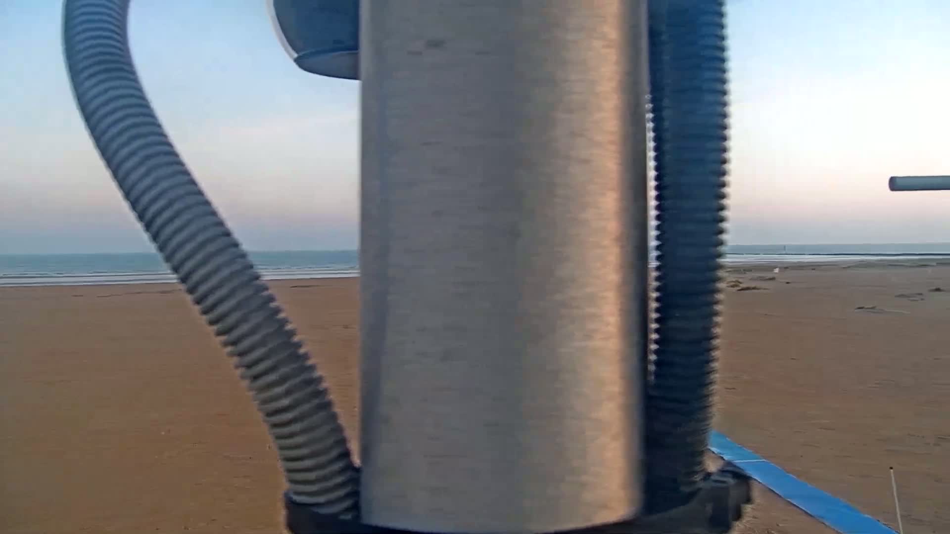 A view through a metal pole shows a sandy beach and calm ocean under a clear sky.