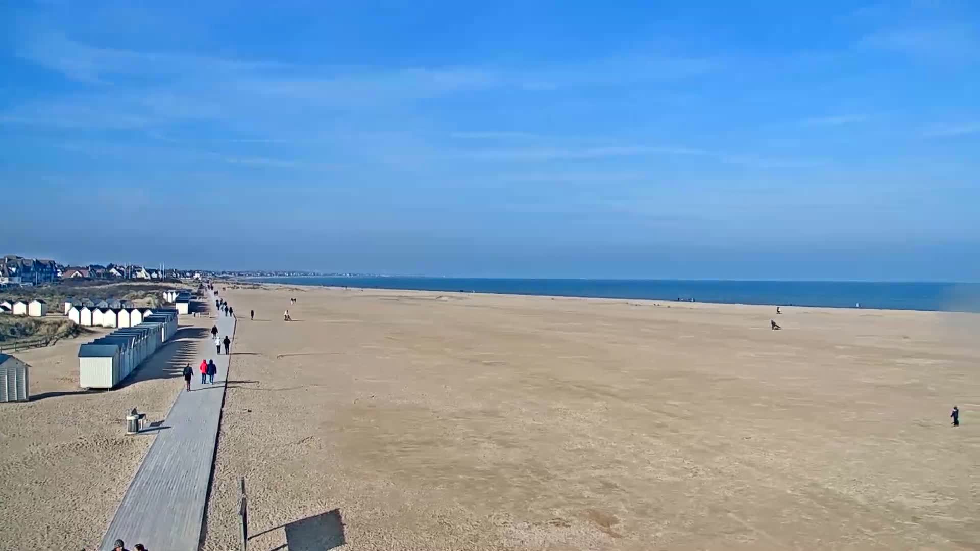 A wide sandy beach stretches to a calm ocean under a clear blue sky, with a boardwalk lined with beach huts and several people walking along it.