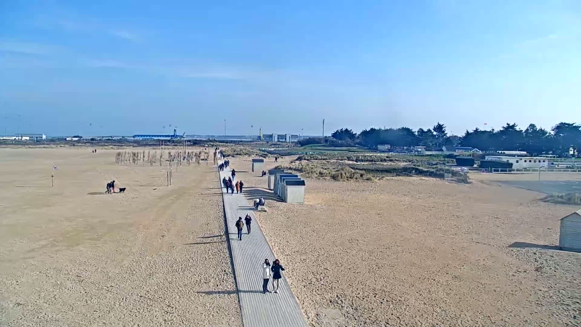A sandy beach on a sunny day features a boardwalk with people walking along it, beach huts, and distant buildings.