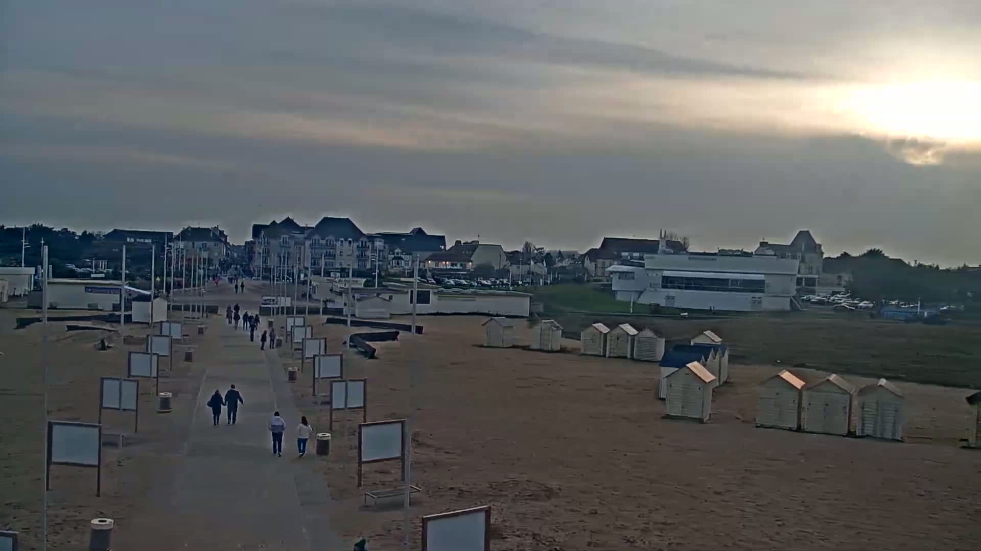 A mostly cloudy, sunset sky illuminates a beach scene with a paved walkway leading to buildings and beach huts.