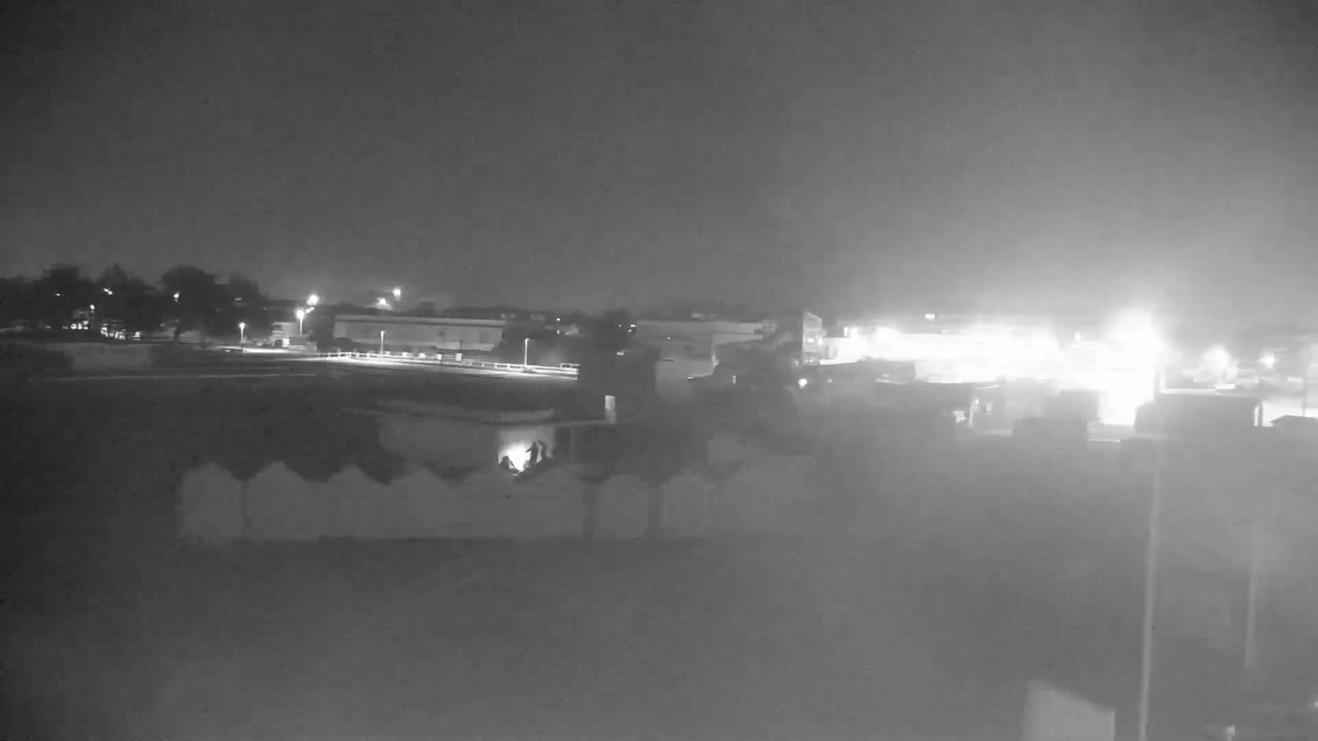 A nighttime, low-light view shows a flat expanse of land with some buildings and trees in the distance, under a hazy sky.