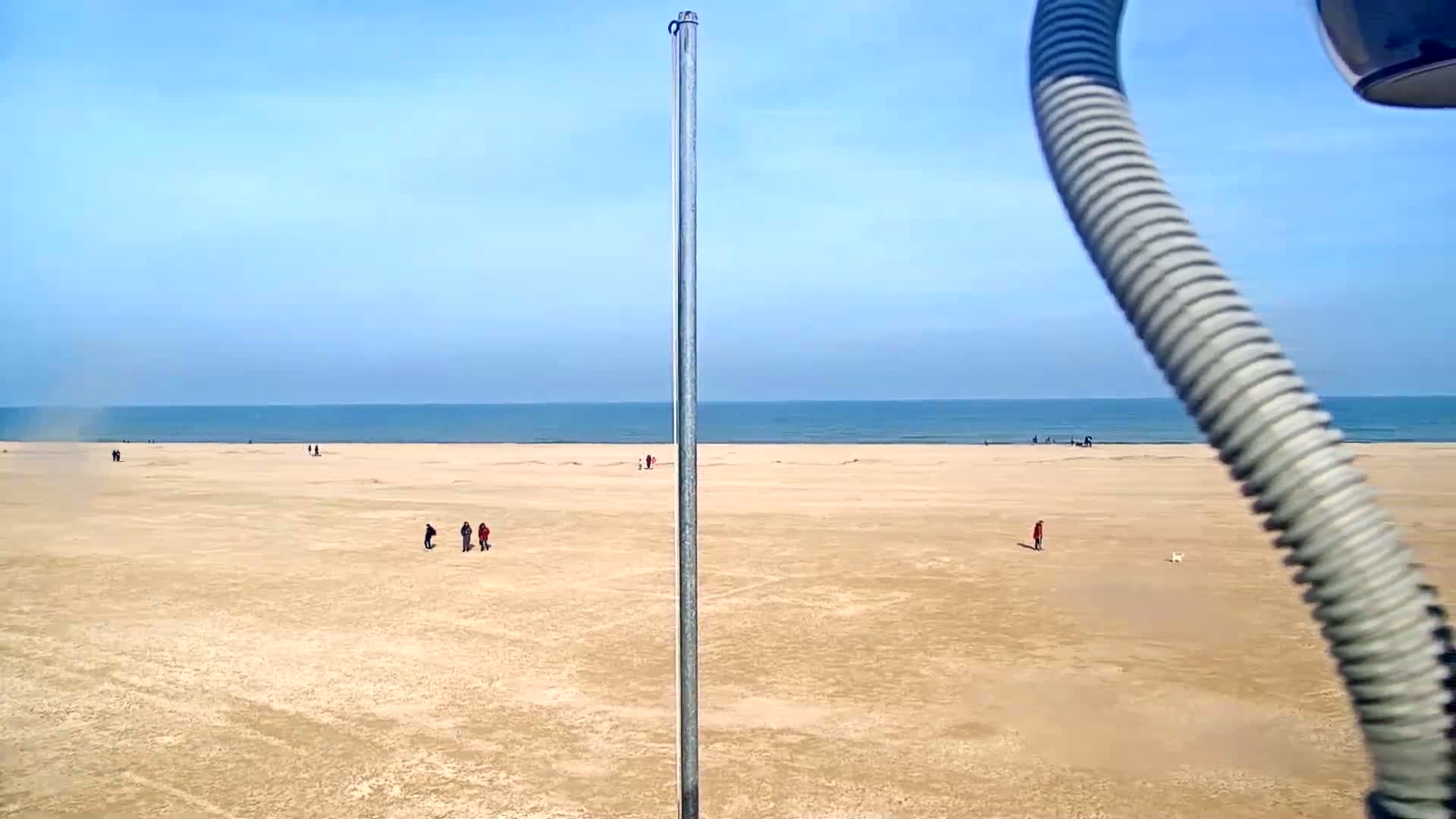 A mostly empty sandy beach stretches to a calm blue ocean under a clear blue sky, with a few people scattered across the sand.
