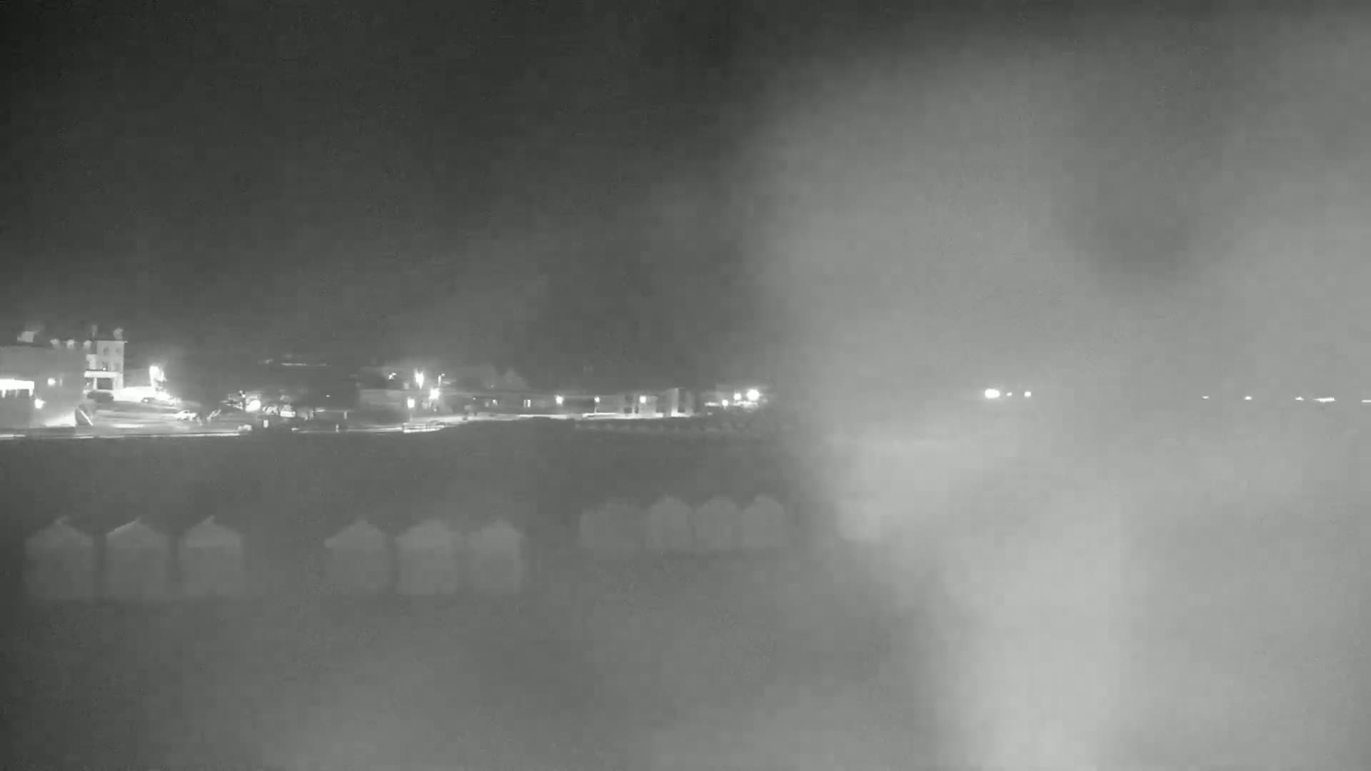 A nighttime beach scene is shrouded in thick fog, with dimly lit buildings and beach huts visible in the distance.