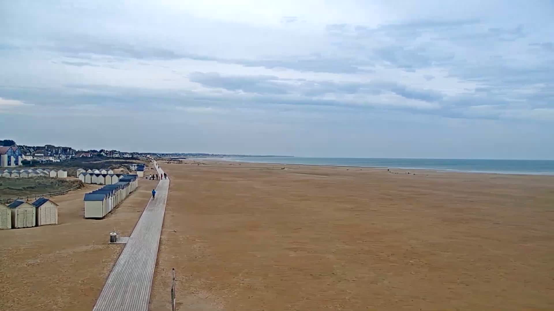 A long, wide sandy beach stretches to a calm ocean under a mostly cloudy sky, with a boardwalk lined with small beach huts leading from a distant town.