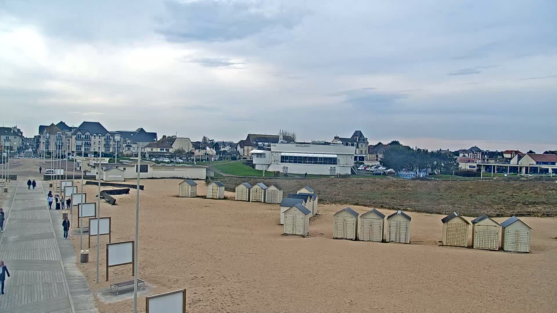 A sandy beach with rows of small beach huts, a boardwalk with people walking, and buildings in the background under a cloudy sky.